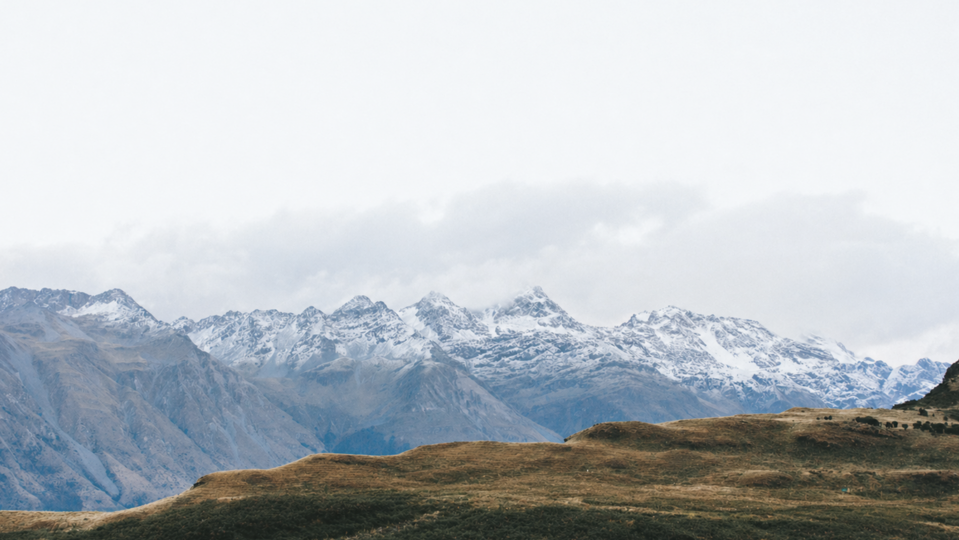 Snow-capped mountains under a cloudy sky with rolling grassy hills in the foreground.