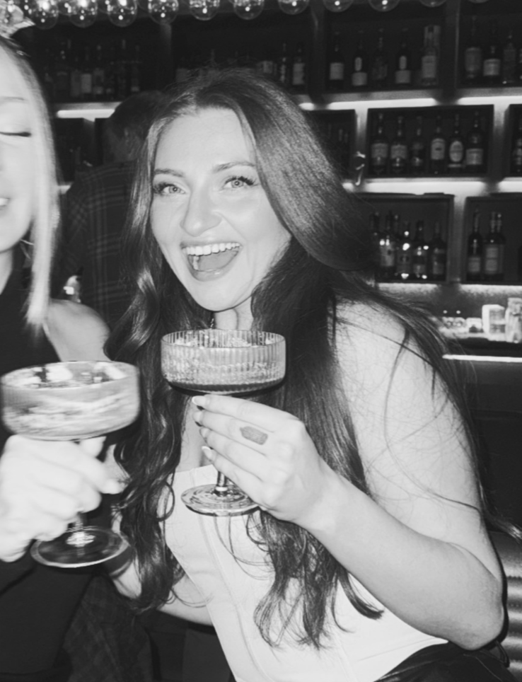 A woman with long dark hair smiling and holding a cocktail glass, in a bar with shelves of liquor bottles in the background. Part of another person is visible on the left.