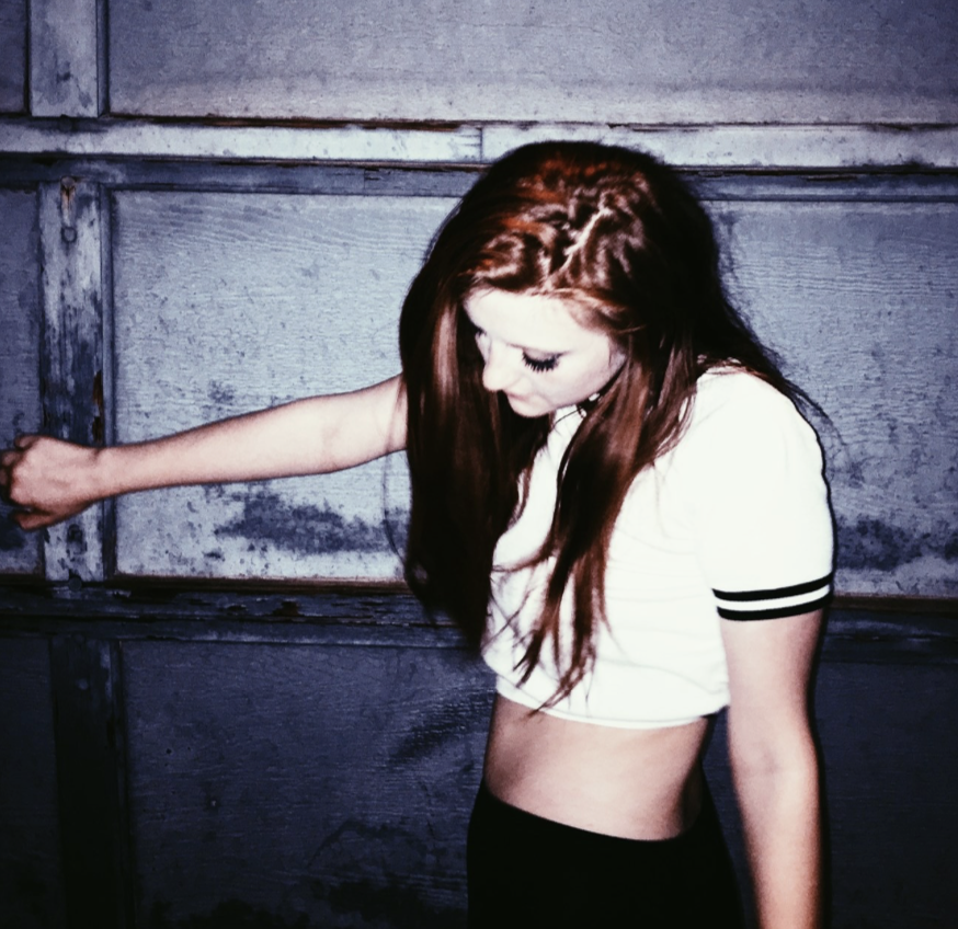 A woman with long red hair and light skin in a white crop top with black stripes on sleeves standing against a weathered wooden wall.