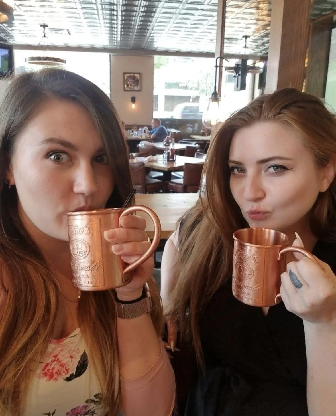 Two women with long hair drinking from copper mugs inside a restaurant.