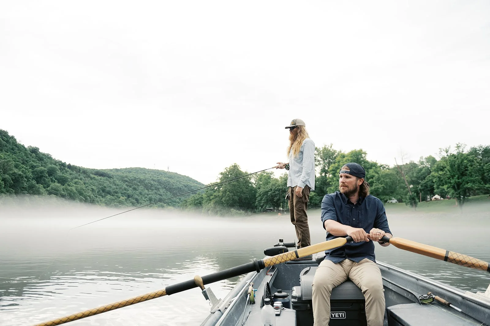 Two men on a boat fishing on a calm lake with mist, surrounded by green hills and trees.