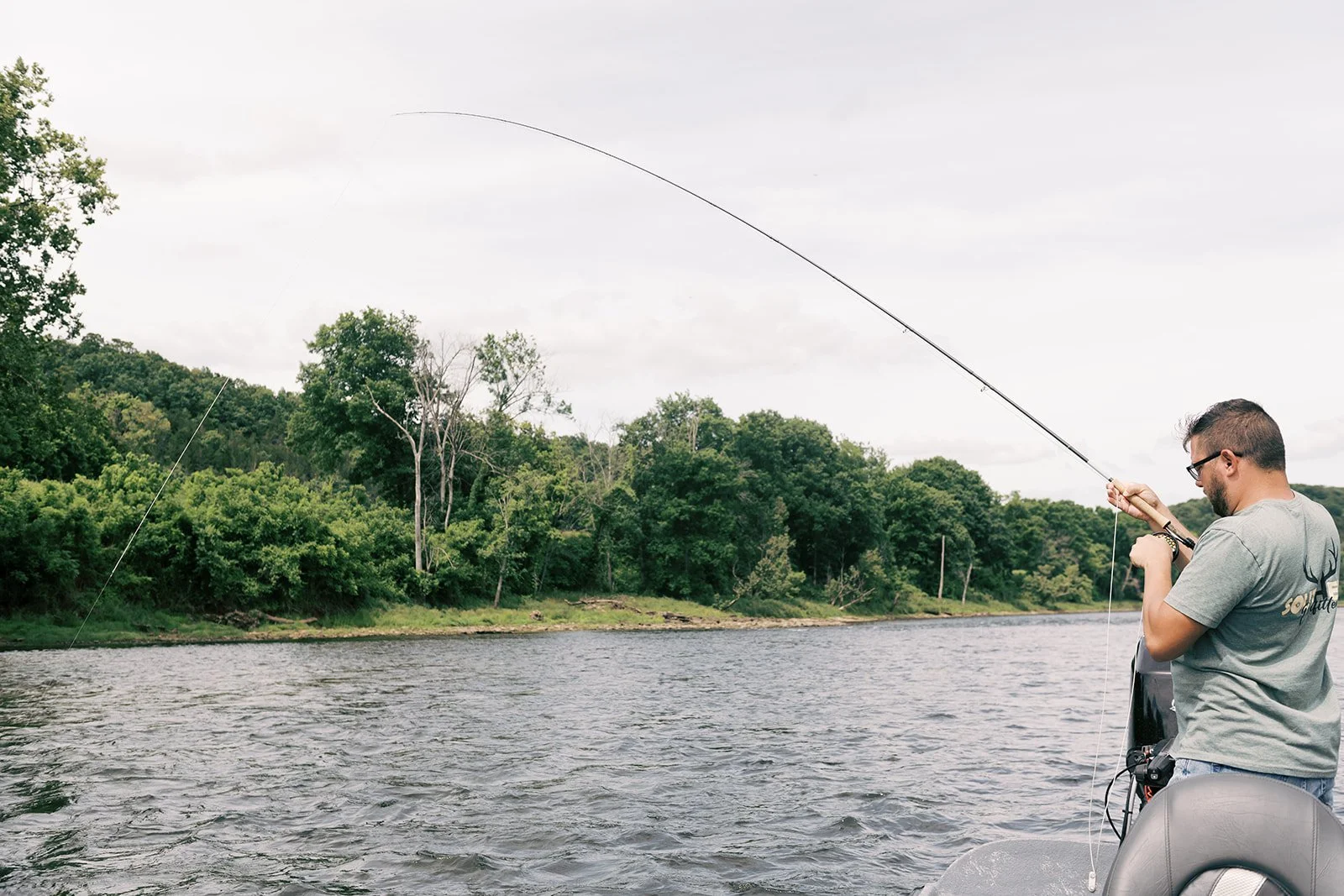 Man fishing from a boat on a river with green trees and cloudy sky in the background.