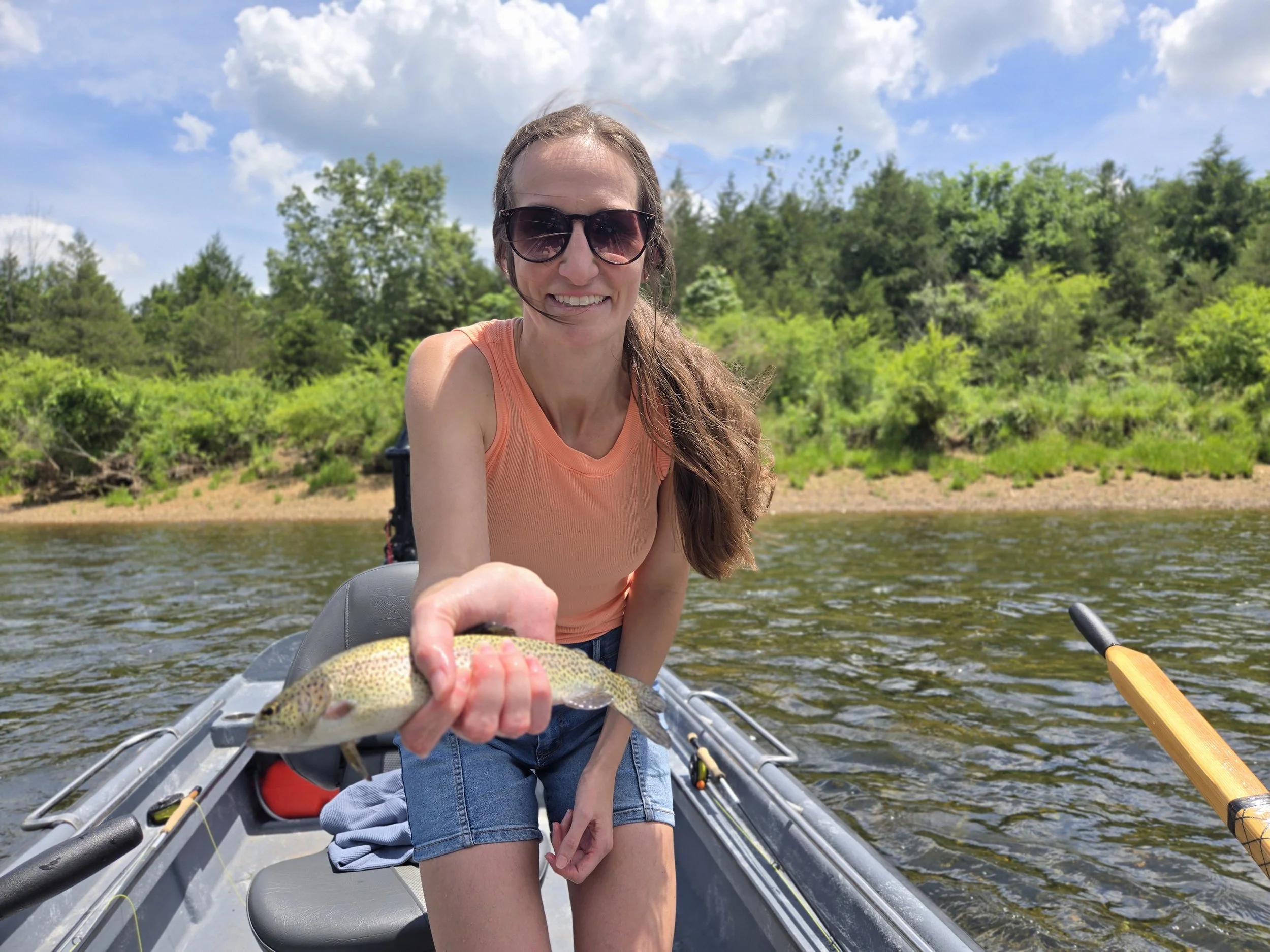 Woman in an orange tank top and sunglasses holding a fish in a boat on a river with green trees and a blue sky in the background.