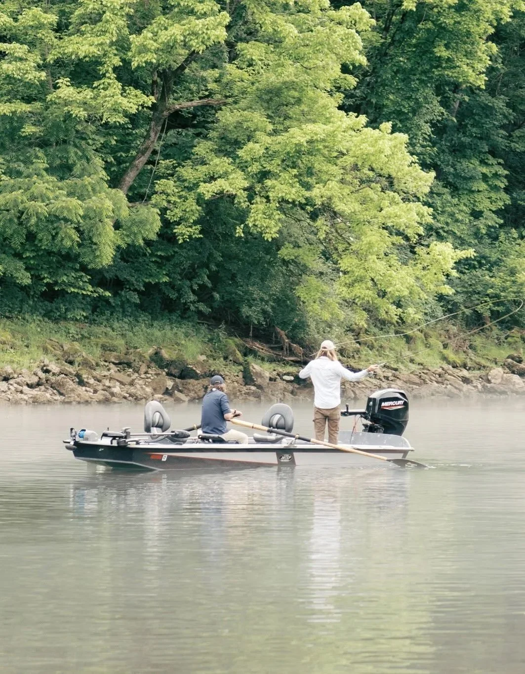 Two men fishing from a boat on a river surrounded by green trees.