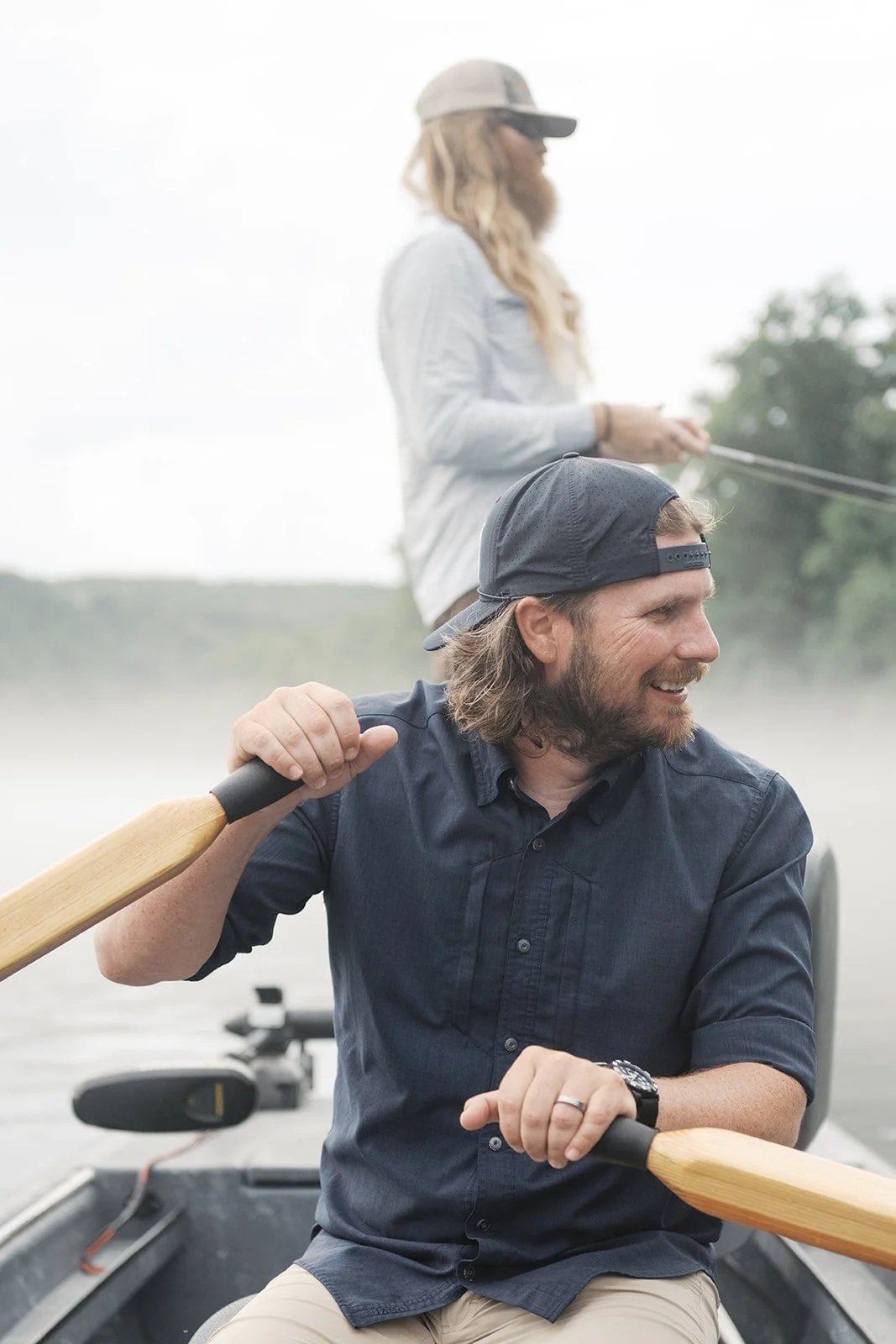 A man with a beard wearing a backward cap and black shirt is smiling and rowing a boat with a wooden oar. A woman with long blonde hair, glasses, and a hat is standing in the background, fishing.