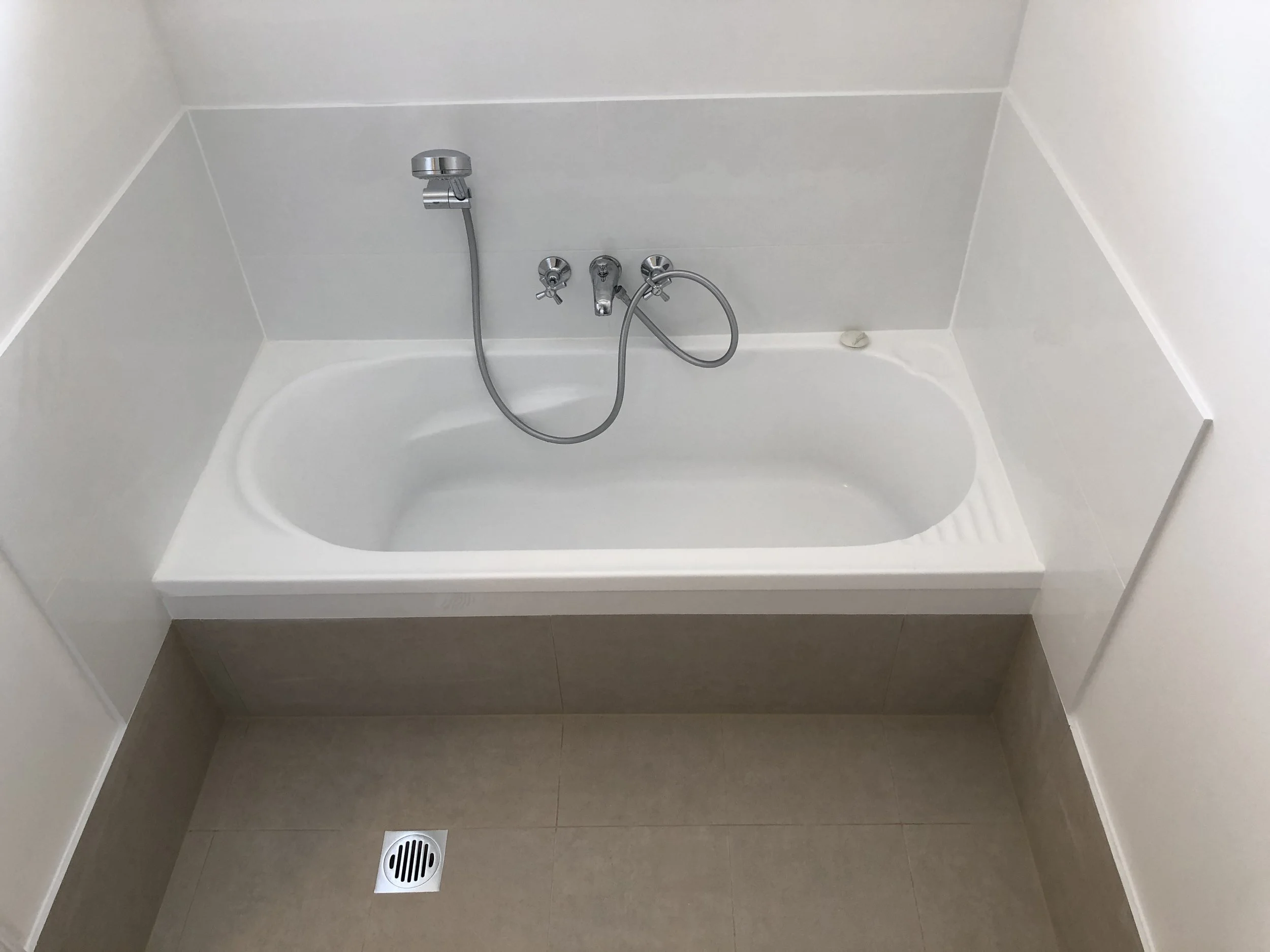 A bathtub with a handheld showerhead and faucet, with a drain and floor drain in a bathroom with light-colored walls and floor.