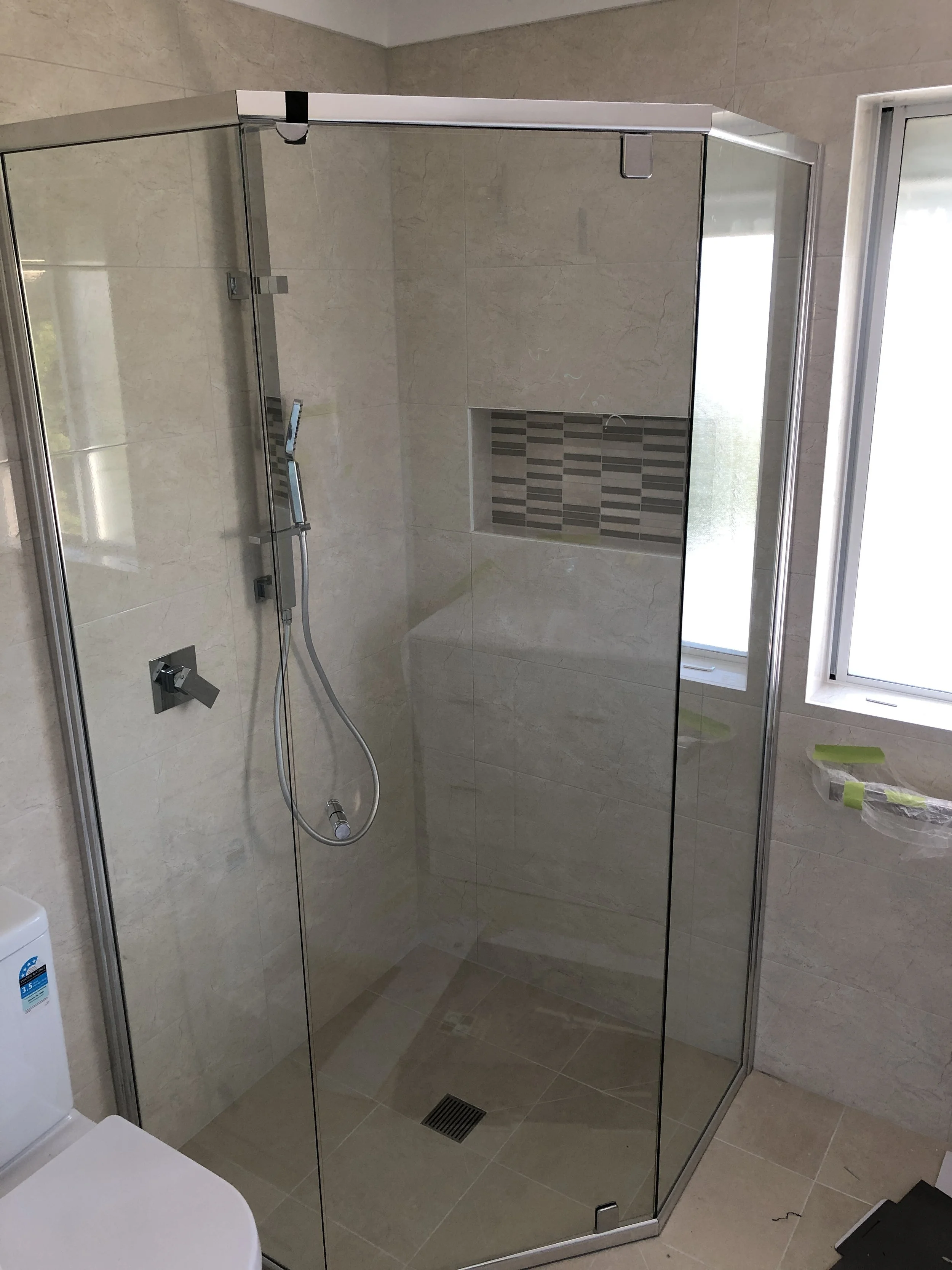 A modern shower with clear glass walls, a built-in tiled niche, a handheld showerhead, and a floor drain, in a beige tiled bathroom.