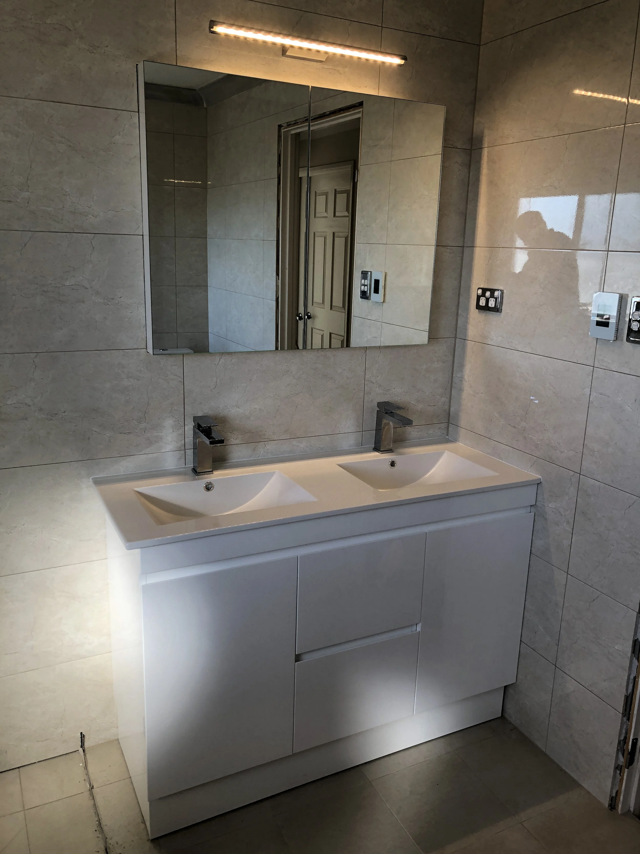 A modern bathroom with a white double sink vanity, a large rectangular mirror above it, and beige ceramic tiles on the walls and floor. Two light switches are visible on the right side of the image.
