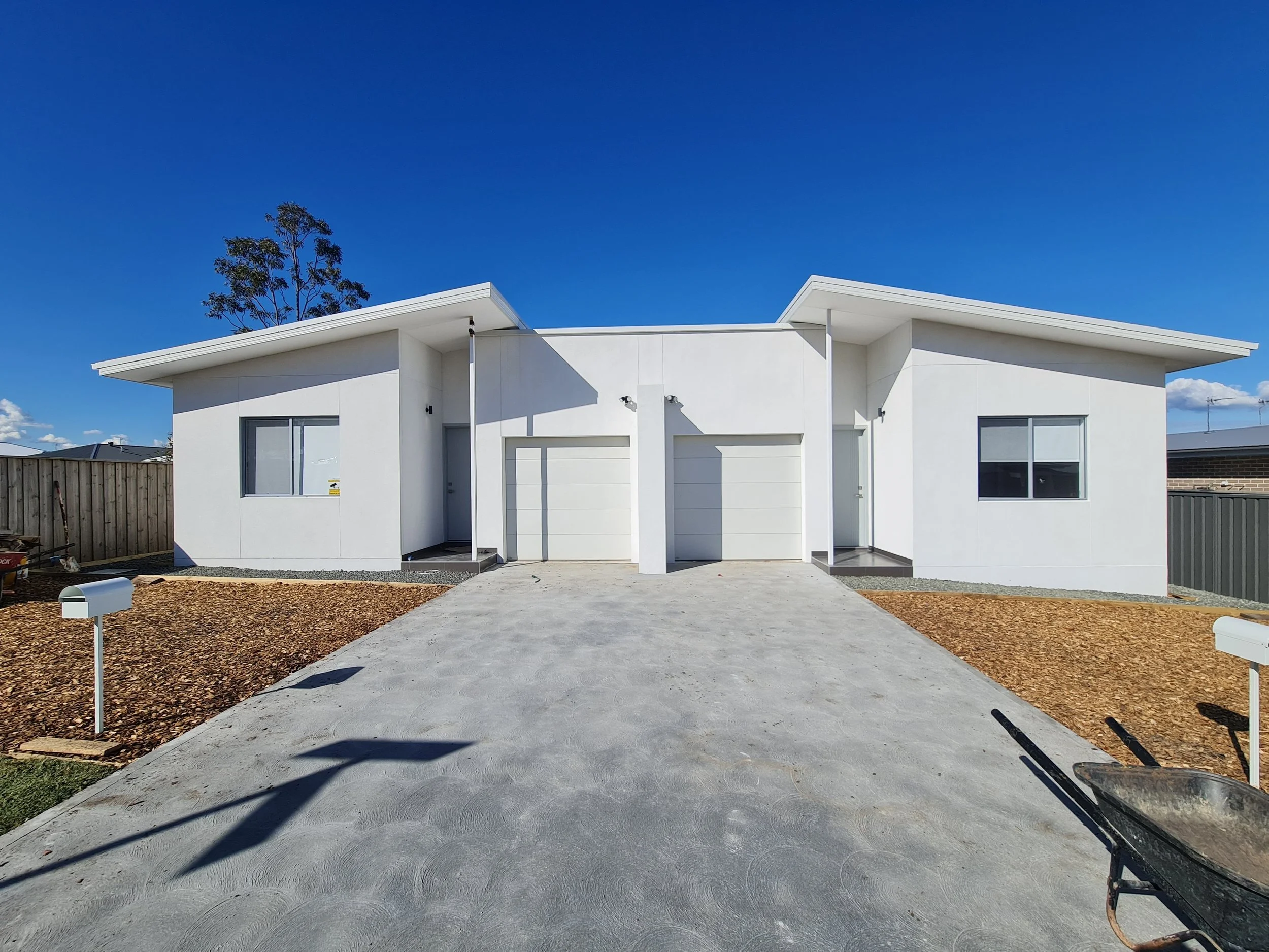 A modern white house with a flat roof and large windows, surrounded by a newly landscaped yard with a concrete walkway, mulch beds, and white mailbox posts, under a clear blue sky.