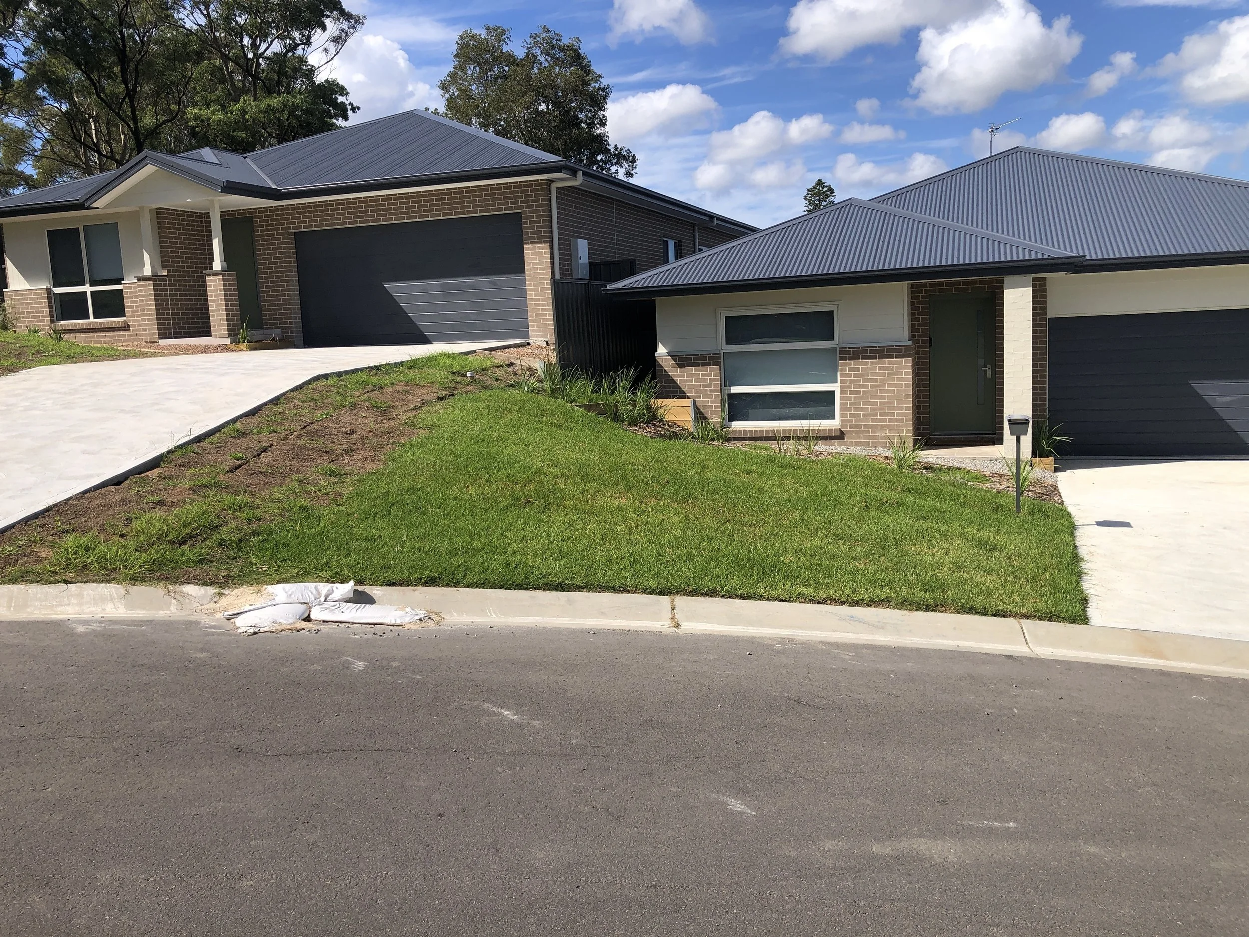 A residential house with a dark metal roof, brick walls, and a garage, surrounded by green grass and a paved sidewalk, with a blue sky and some clouds above.
