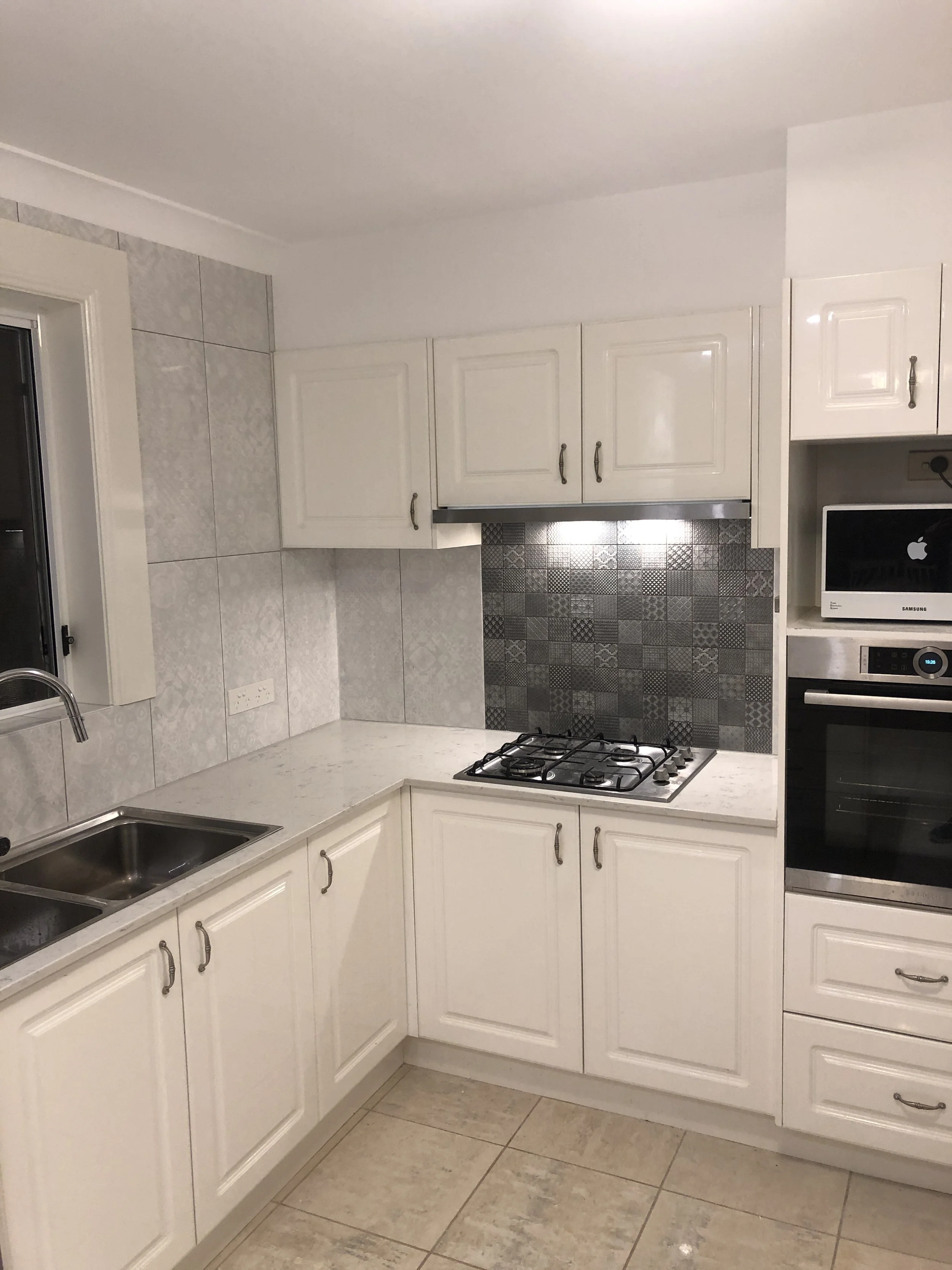 Kitchen with white cabinets, gray tiled backsplash, gray countertop, stainless steel sink, and built-in stove. There is also a built-in oven and microwave, and a window with a black frame.
