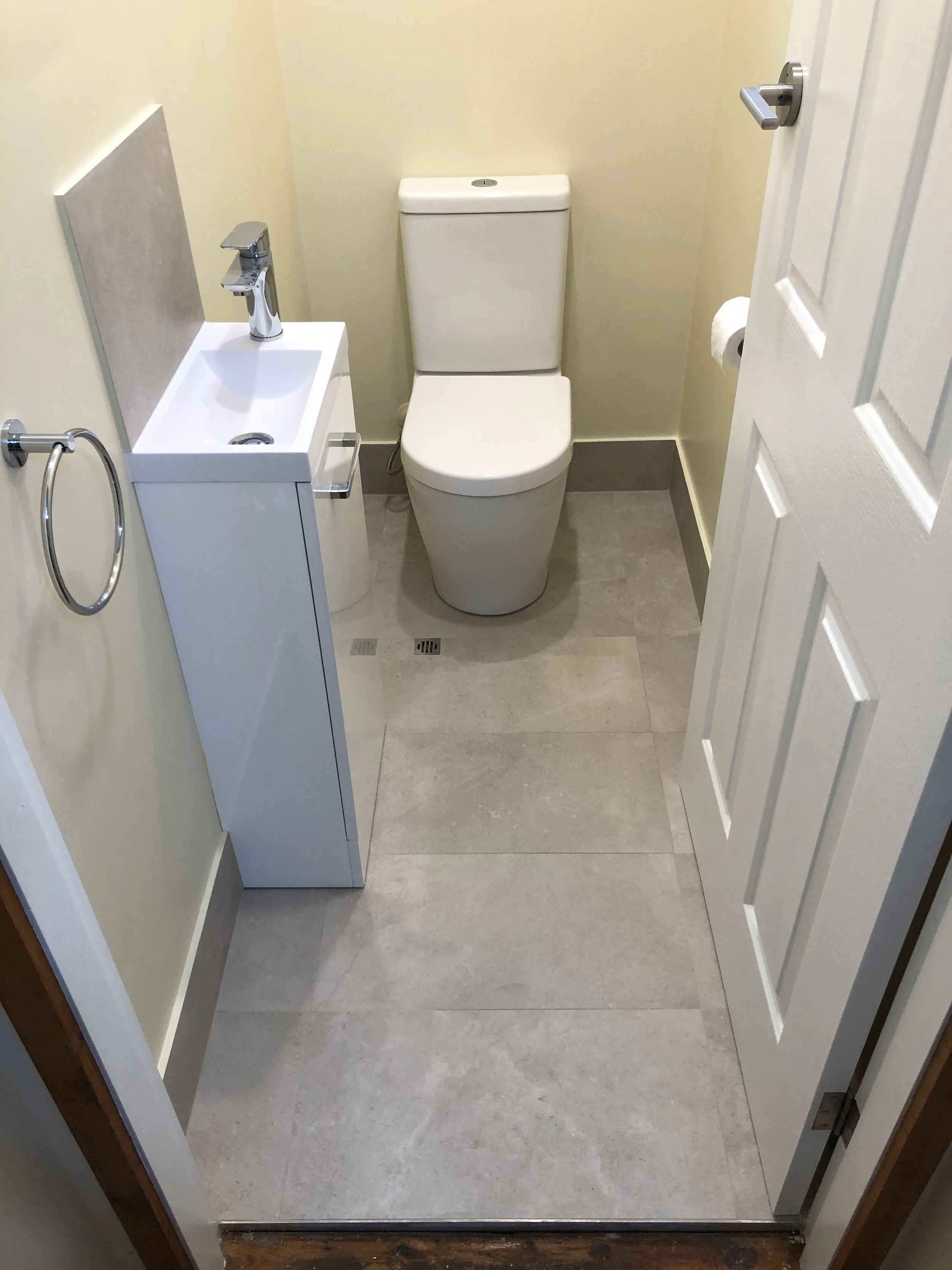Small bathroom with a white toilet and a small white sink with a chrome faucet, beige walls, light gray tile floor, and a white door open outward.