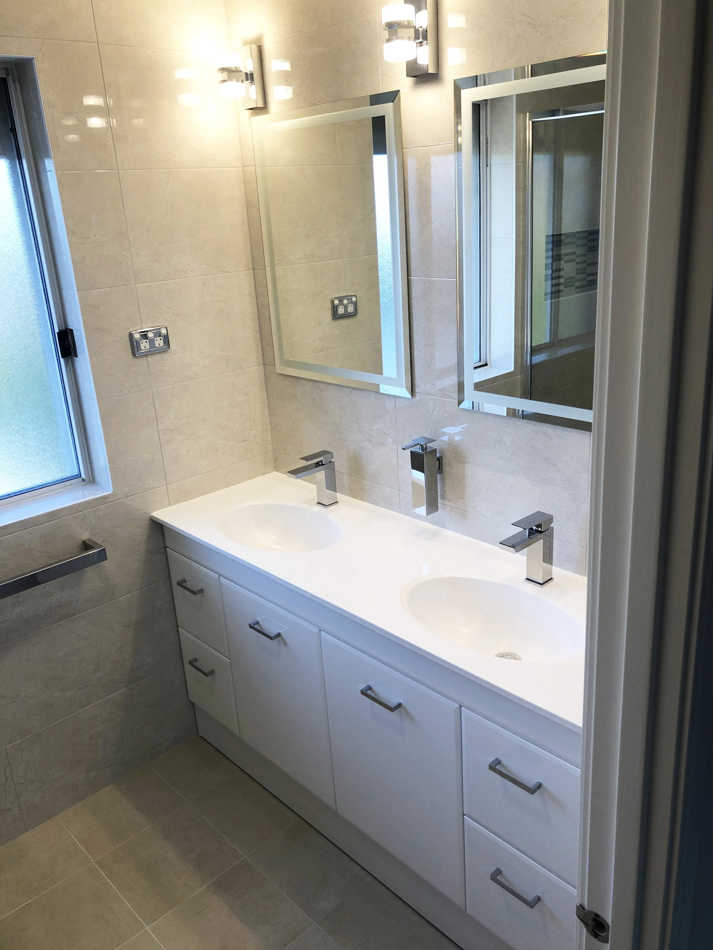 Bathroom with two sinks, mirrors, and beige tiled walls and floor, with a window on the left side.