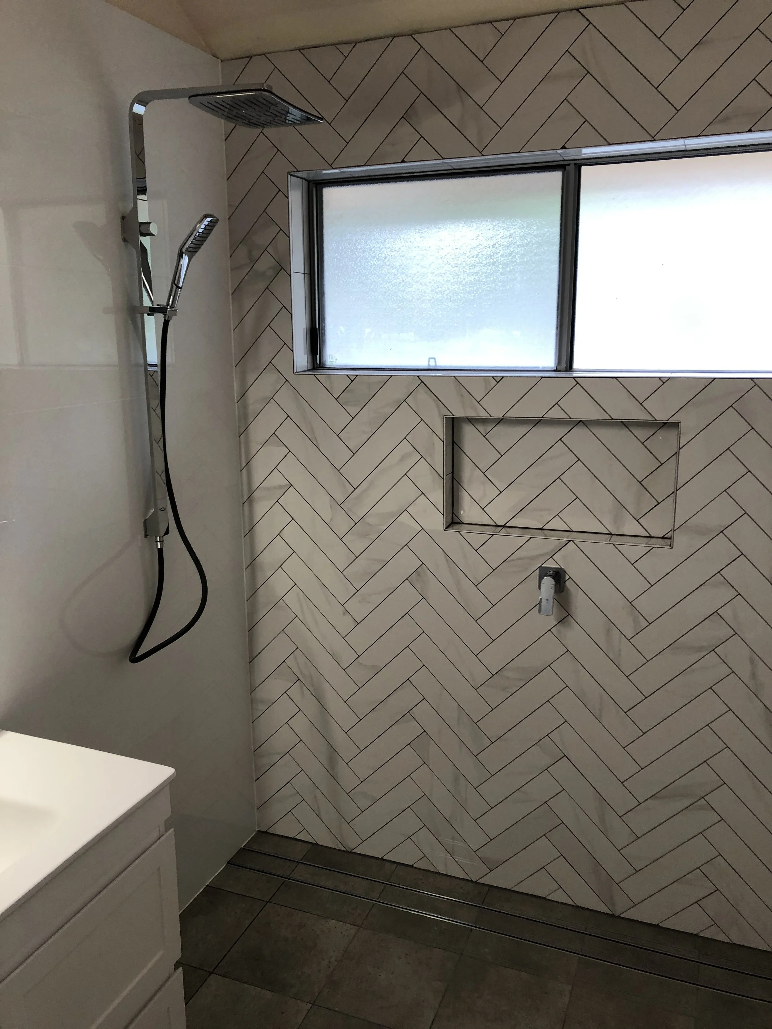 A modern bathroom shower area with beige herringbone-patterned wall tiles, a frosted window, a built-in niche, and a chrome showerhead with a handheld spray.