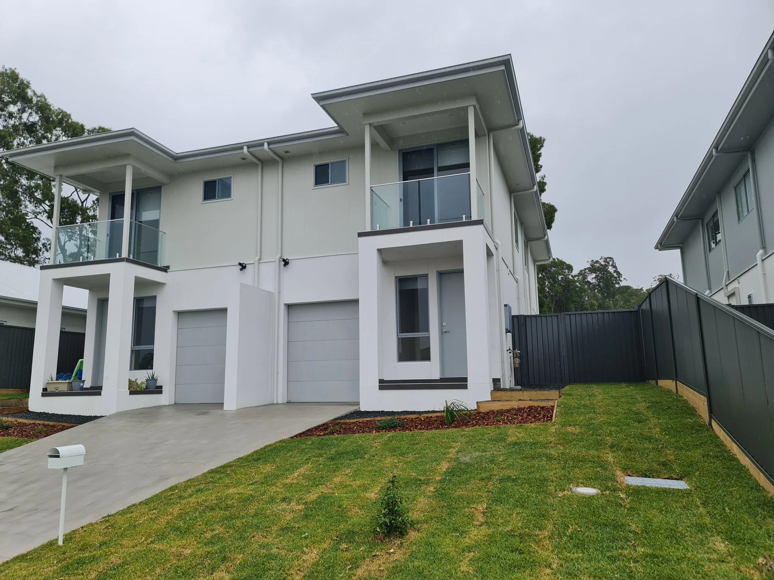 Modern two-story house with white exterior, large windows, a glass balcony on the second floor, and a driveway in front. The yard has green grass and small plants, with a cloudy sky overhead.