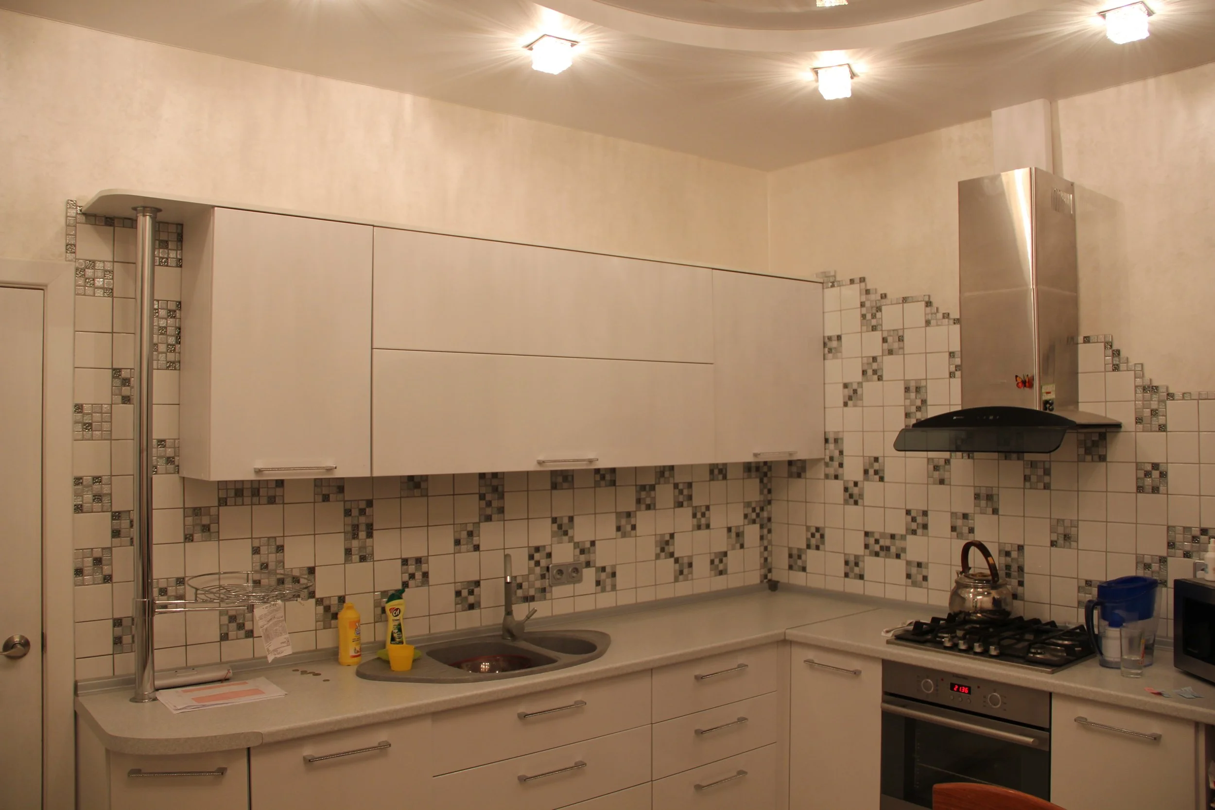 A kitchen with white cabinets, a tiled backsplash with a geometric pattern, a kitchen faucet, a small sink, a countertop with cleaning supplies, a stove with a kettle, and overhead lighting fixtures.