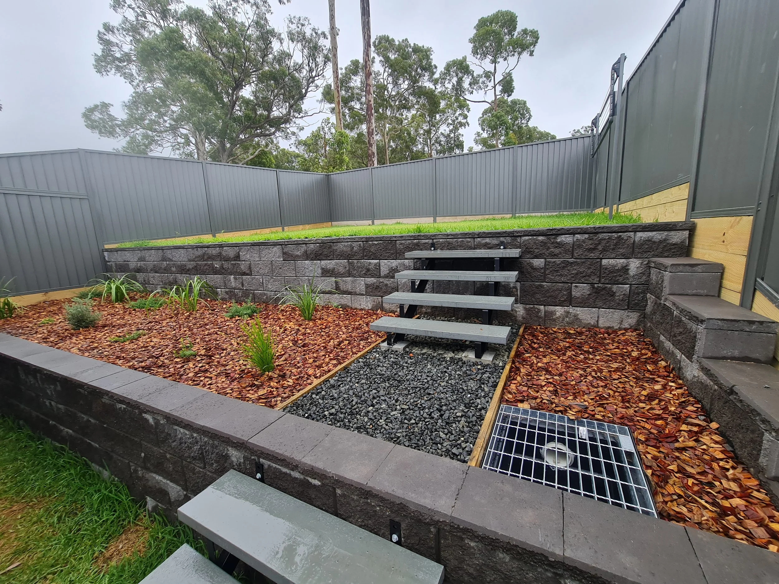 A backyard garden with a raised bed filled with plants, a retaining wall made of dark gray bricks, and a tiered pathway with gray steps. There is a tall gray fence surrounding the yard and a partly cloudy sky overhead.