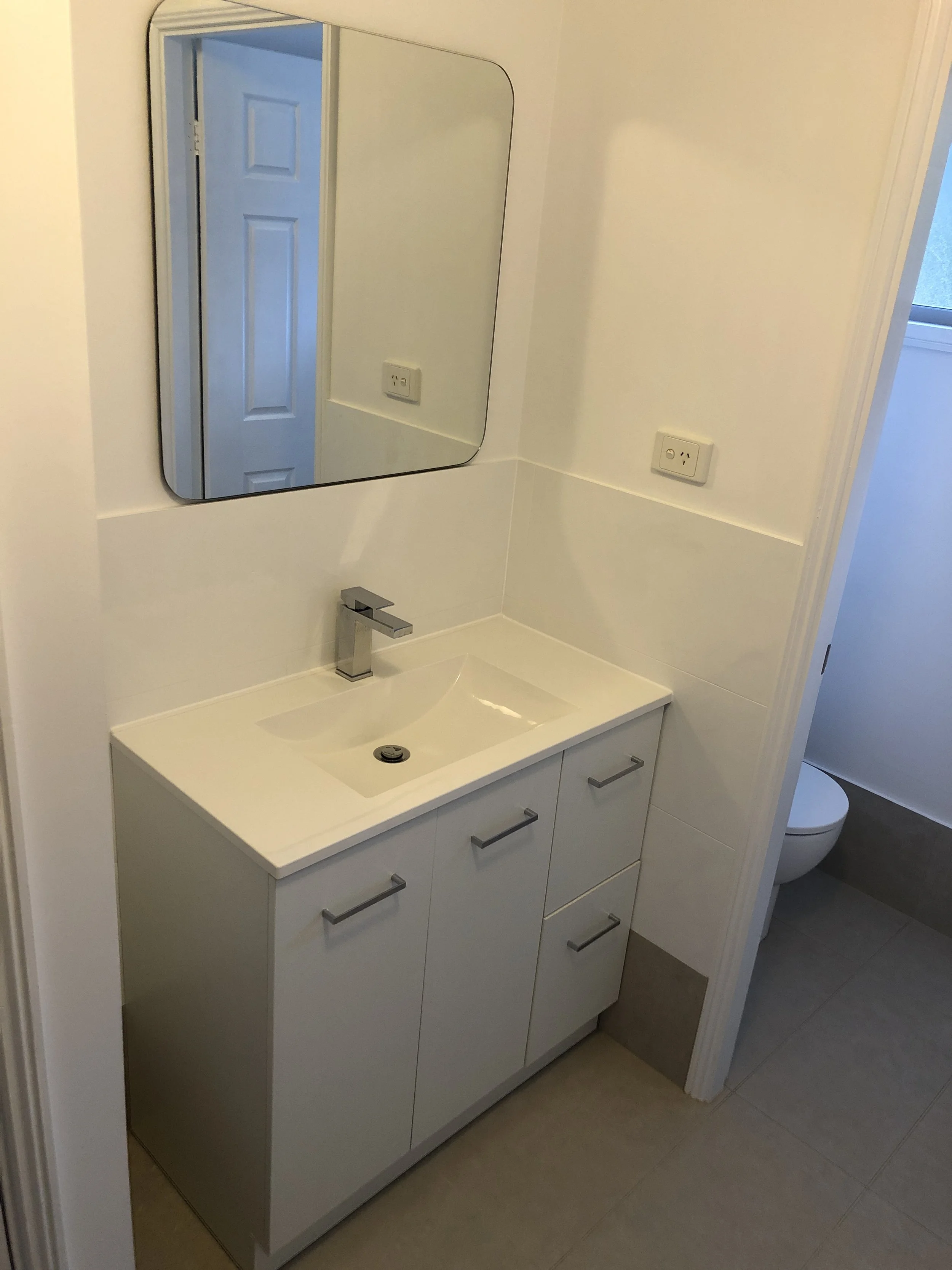 Bathroom vanity with white cabinet, marble countertop, and rectangular mirror above. There is a modern faucet and visible electrical outlets. A toilet is partially visible in the background, and the room has tiled flooring.