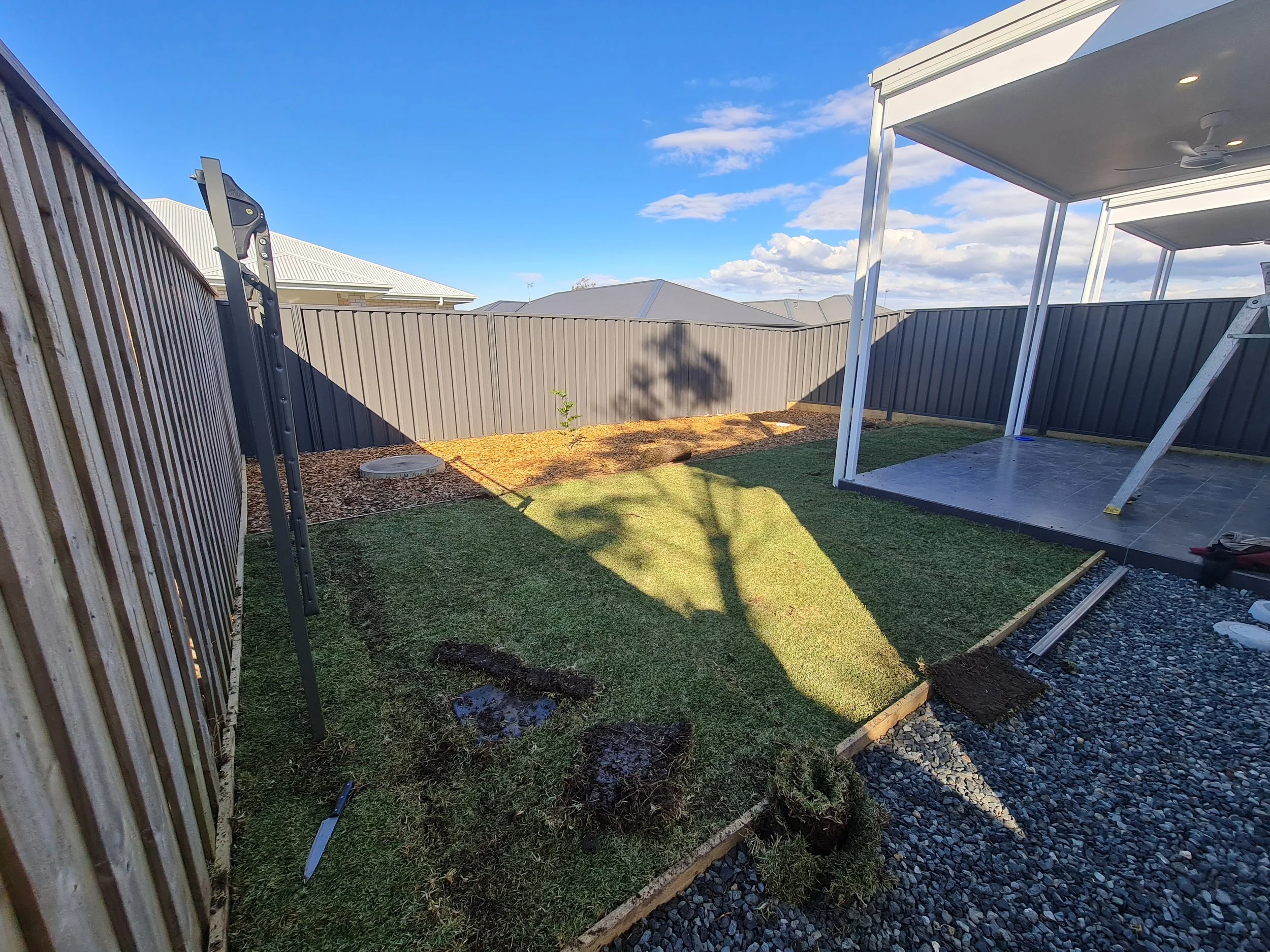 A backyard with a gray metal fence, a small patch of grass, a section of mulch with a small plant, and a covered patio with a ceiling fan. Construction materials and tools are present, indicating ongoing yard or patio work. The sky is blue with some 