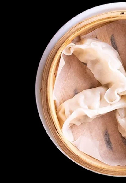 Close-up of uncooked dumplings in a round bamboo steamer with parchment paper lining, on a black background.
