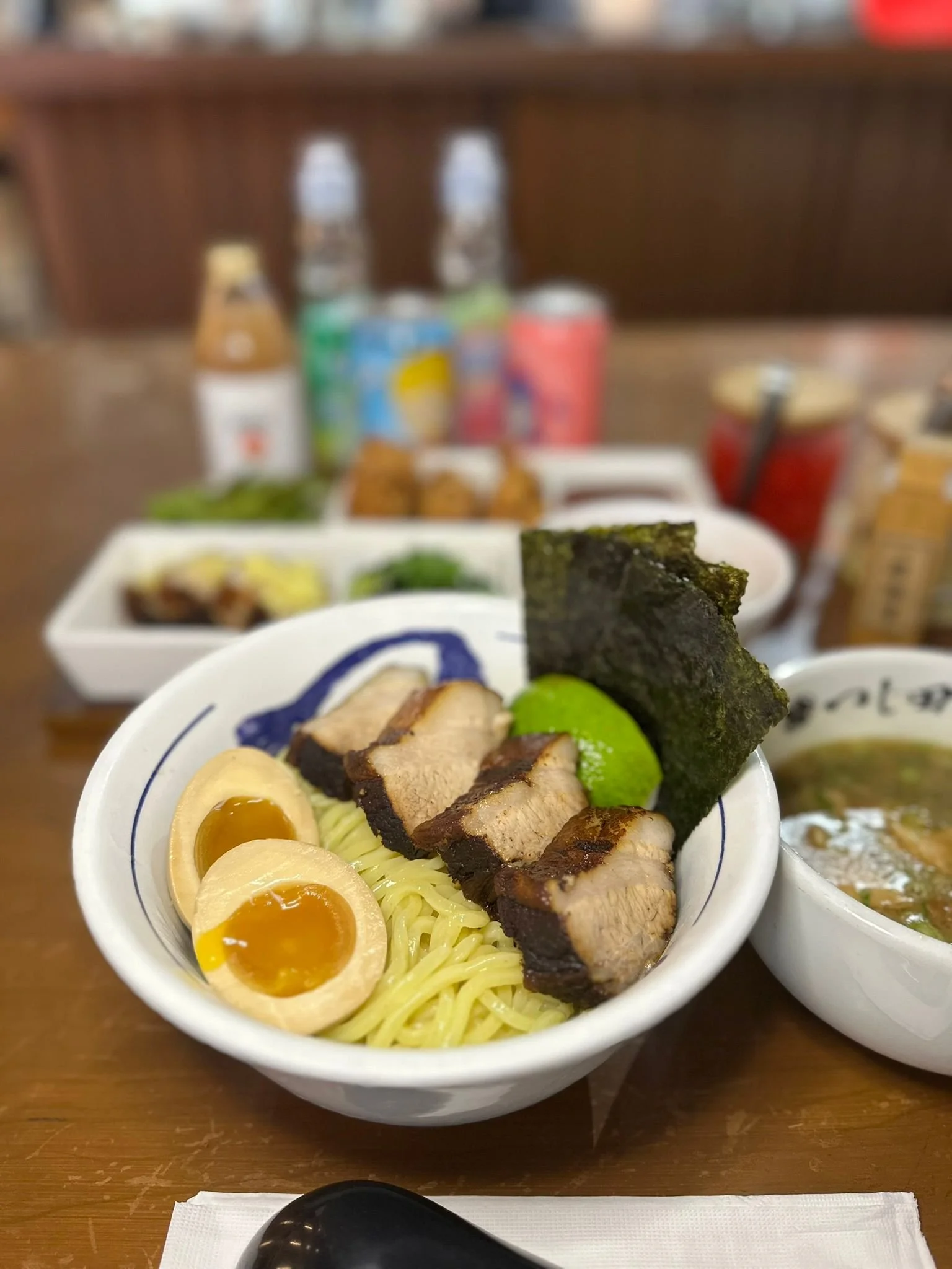 A bowl of ramen with sliced pork, soft boiled eggs, green onions, and seaweed, with a side of ramen toppings and drinks in the background.