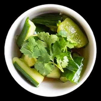A small white bowl filled with chopped cucumber, cilantro leaves, and lime wedges on top.