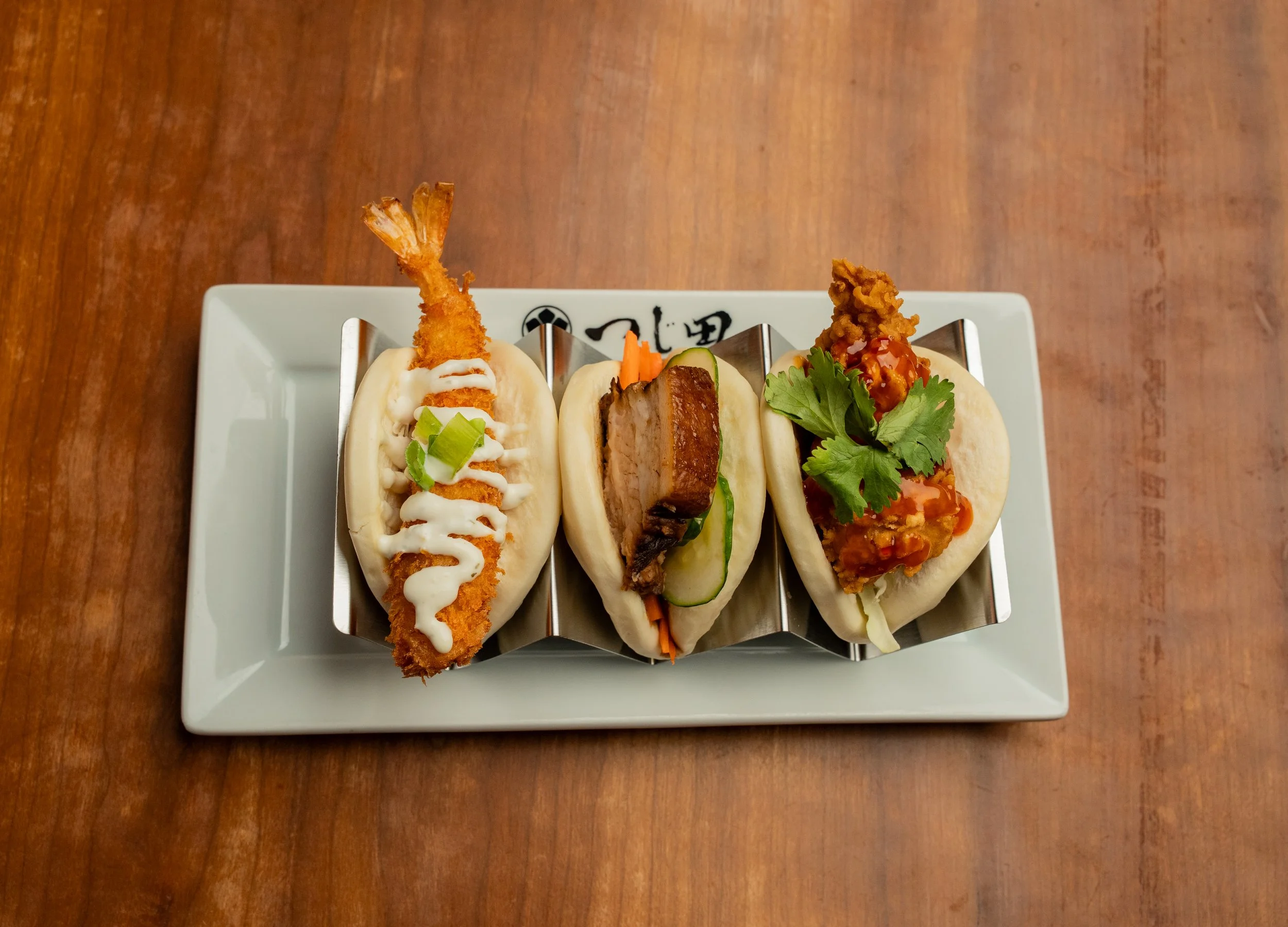 Three Asian-style sandwiches with different fillings on a white plate, garnished with vegetables and sauces, placed on a wooden table.