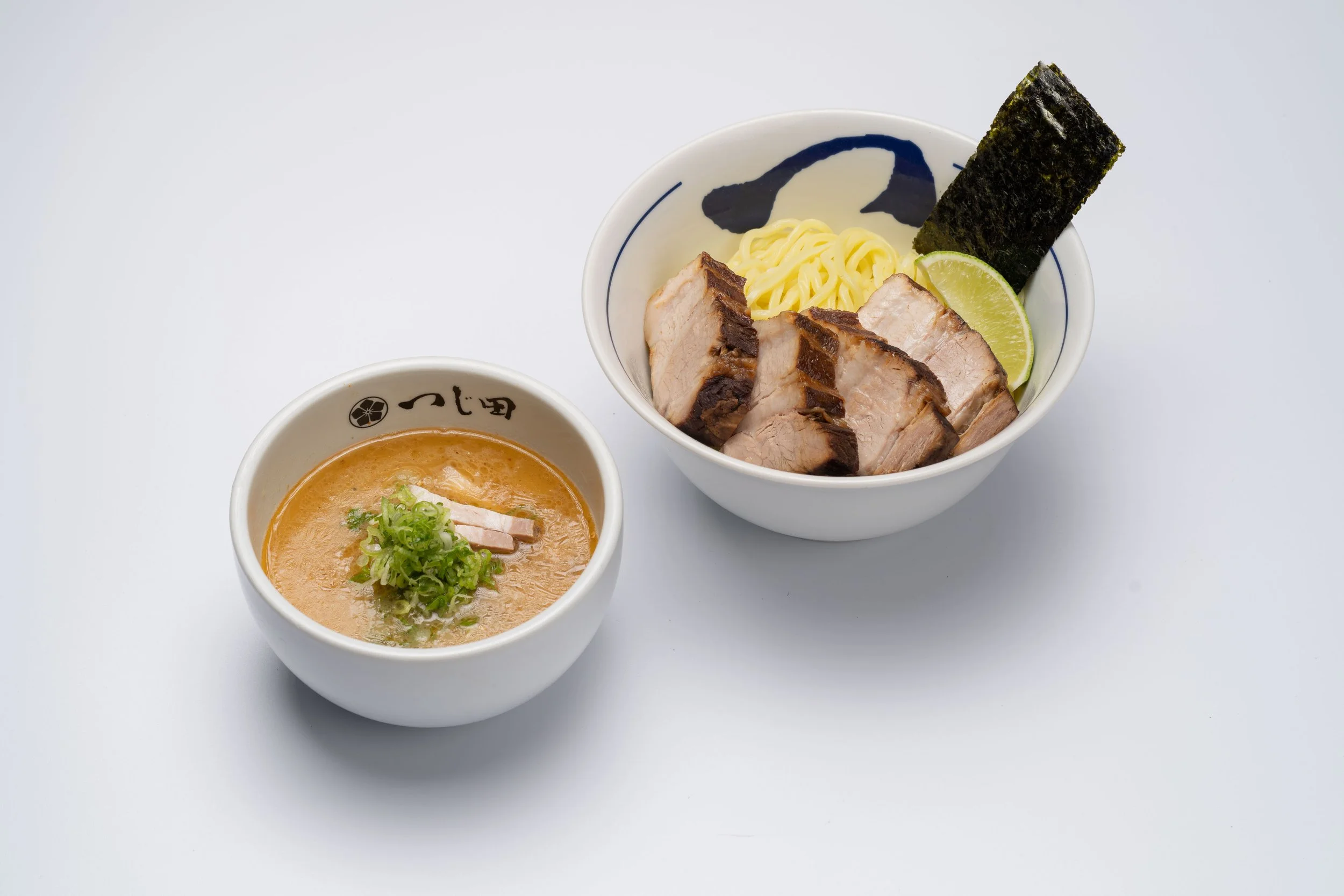 A bowl of ramen noodles topped with slices of pork, a piece of nori seaweed, and a lime wedge. Next to it is a smaller bowl of creamy, orange-colored soup garnished with green onions.
