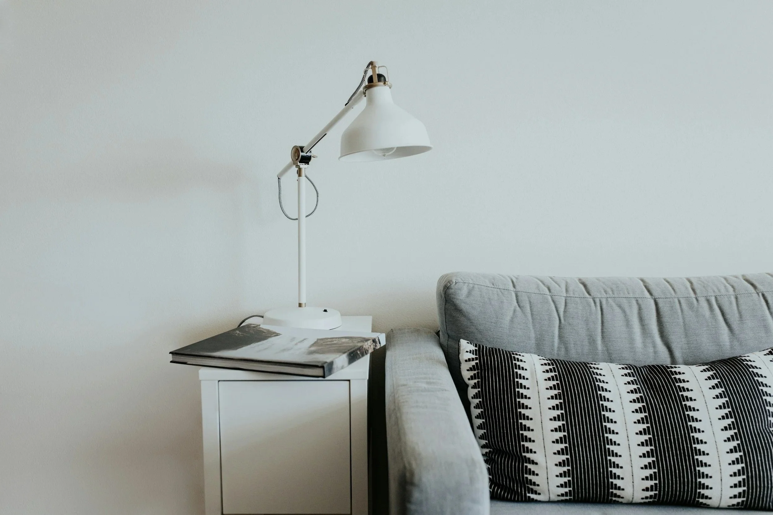 A white adjustable desk lamp on a white nightstand next to a gray couch with black and white patterned pillows, in a room with plain white walls.