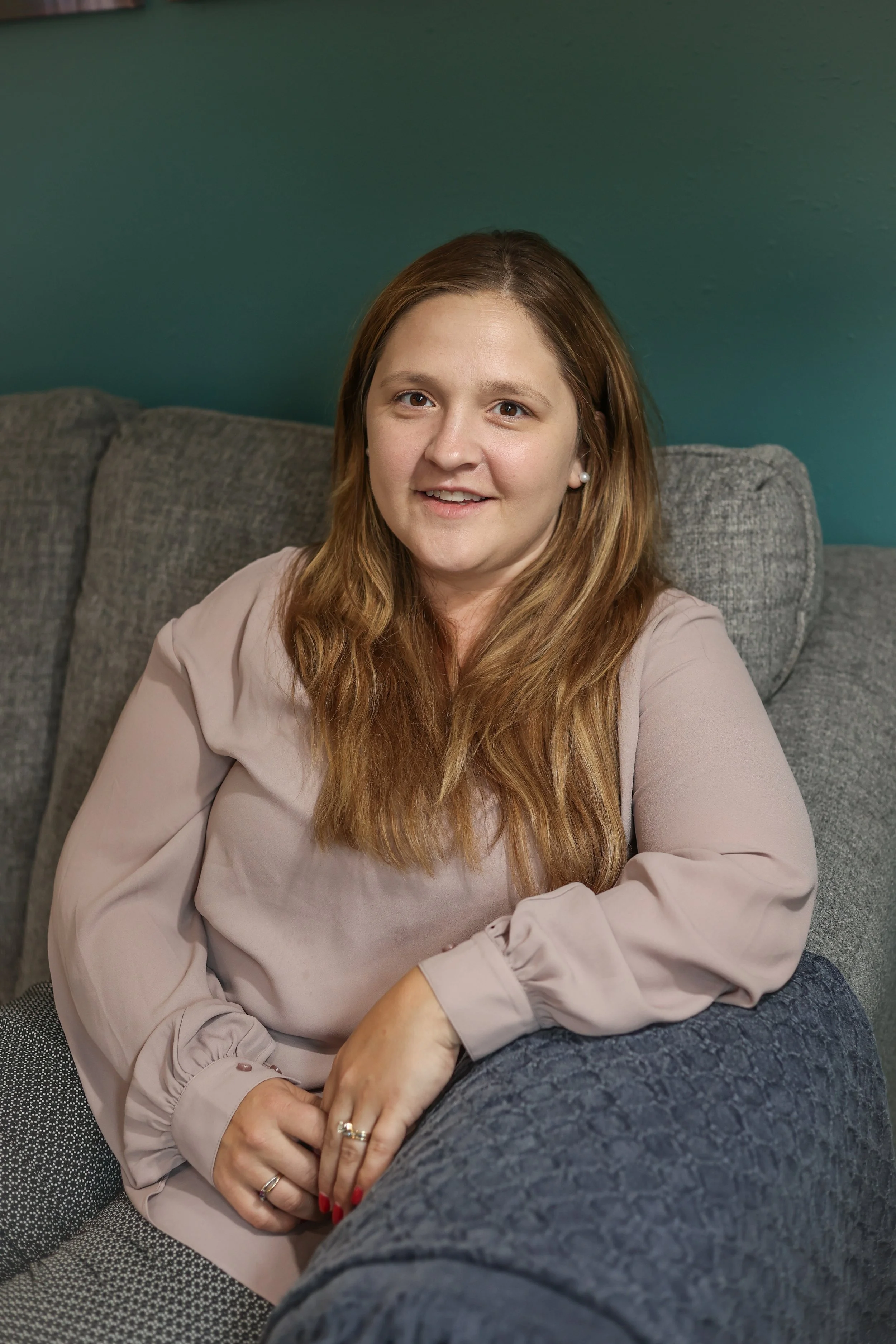 A woman with long, wavy, light brown hair sitting on a gray couch against a teal wall. She is wearing a light beige blouse with puffed sleeves and pearl earrings. She is smiling and looking at the camera.