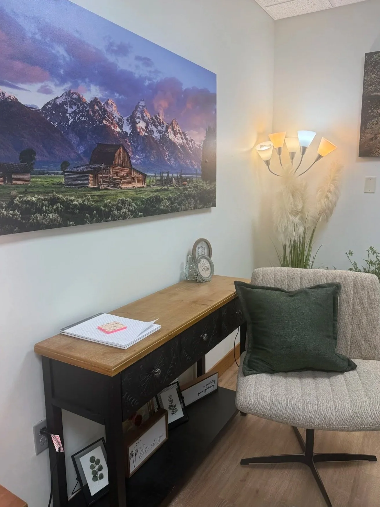 A cozy corner with a framed mountain landscape photo hanging on the wall, a lamp with multiple shades, a large pampas grass plant, a beige armchair with a dark cushion, a wooden table with decorative items and framed pictures underneath, and some notebooks on top.