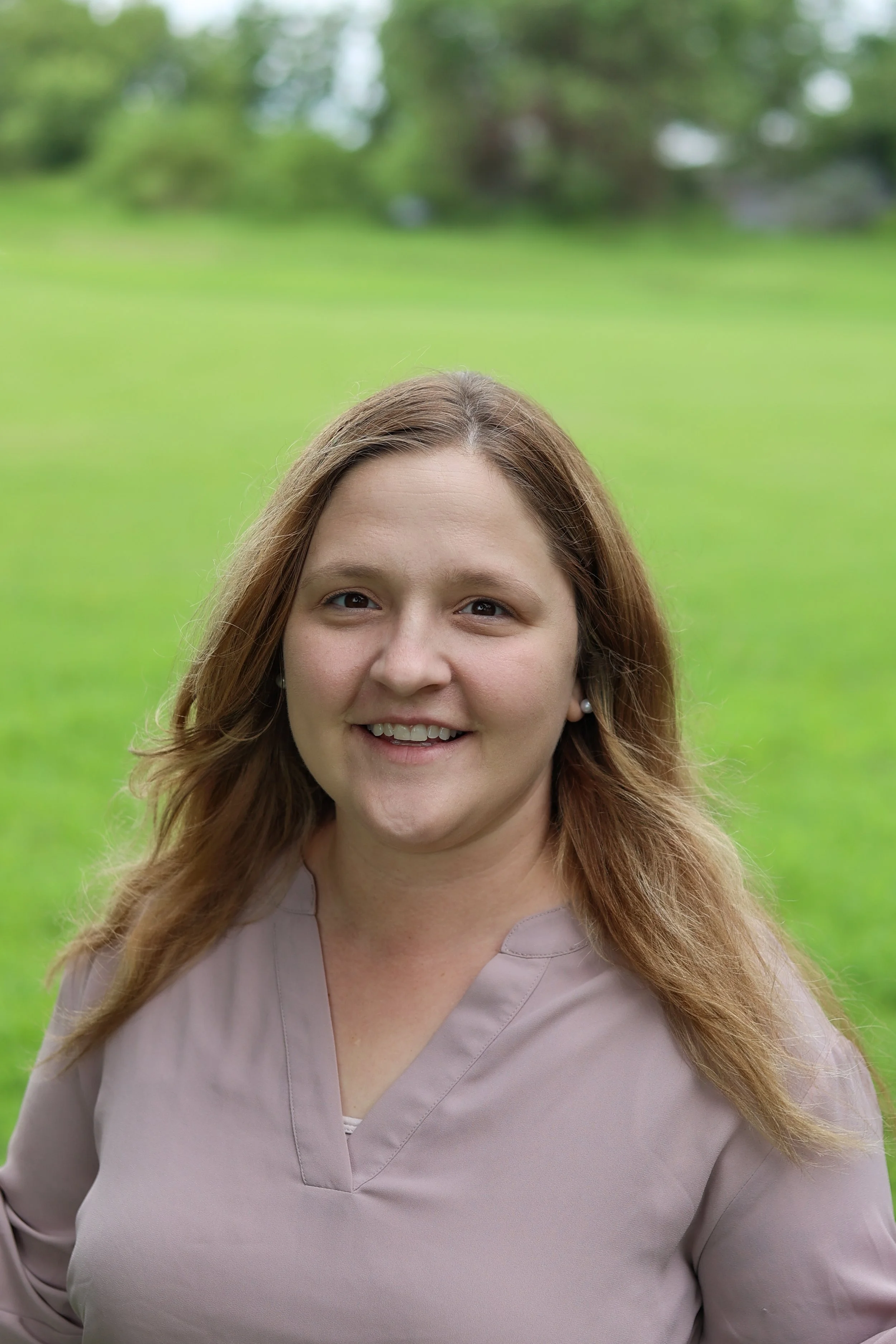 A woman with light skin and long, wavy, reddish-brown hair, smiling outdoors against a green grassy background.