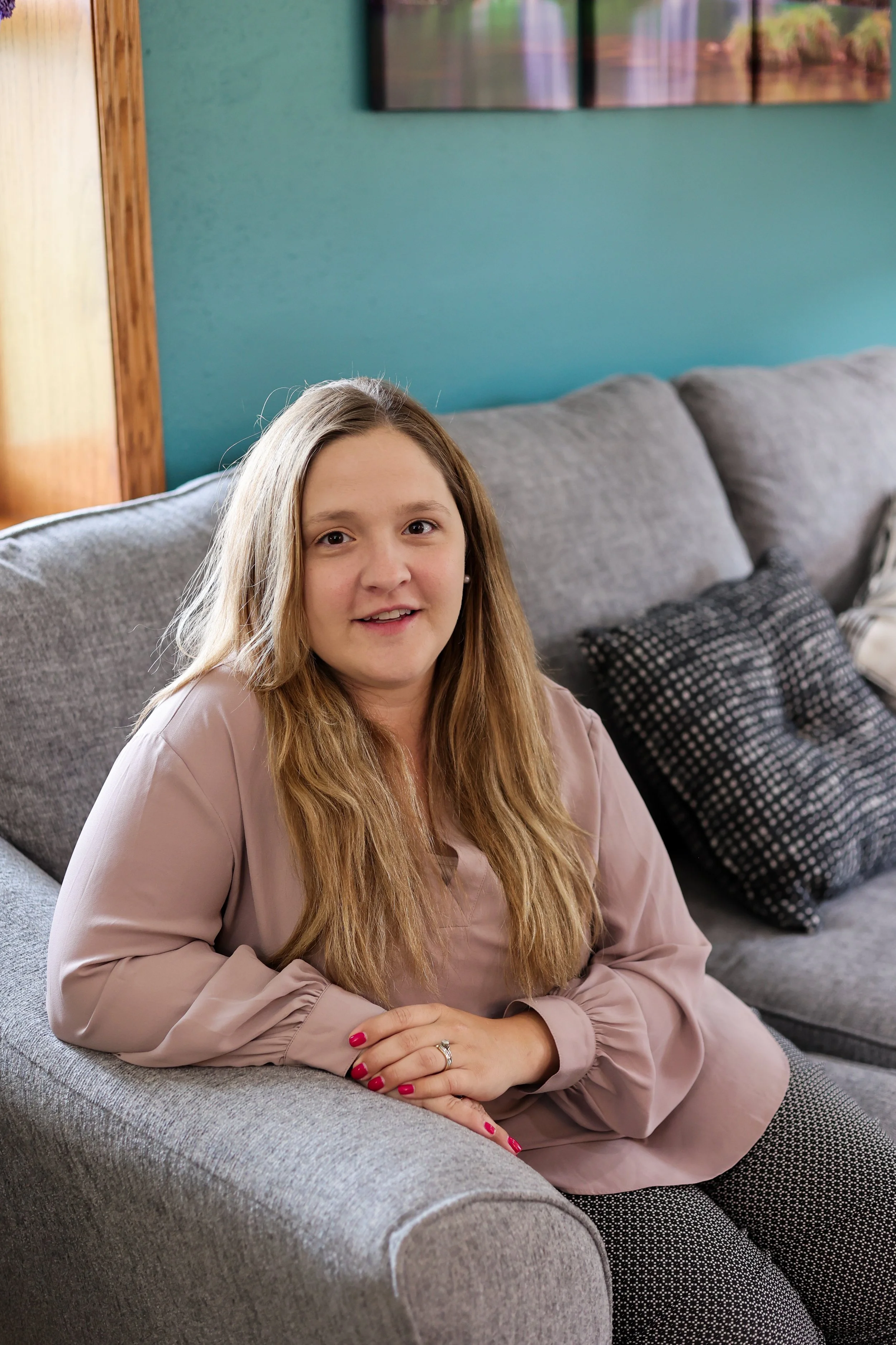 A woman with long blonde hair sitting on a gray couch in a living room with teal walls. She's smiling and has her hands folded, showing a ring on her finger. She is wearing a beige blouse with long sleeves and has bright red nail polish.
