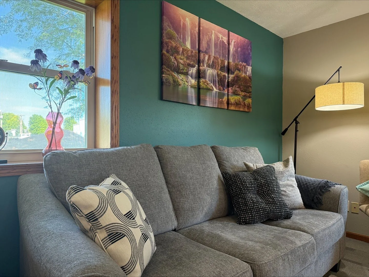 A living room with a grey sofa decorated with three pillows, a colorful painting of waterfalls on a teal wall, a window with a pink vase holding purple flowers, and a standing lamp with a beige shade.