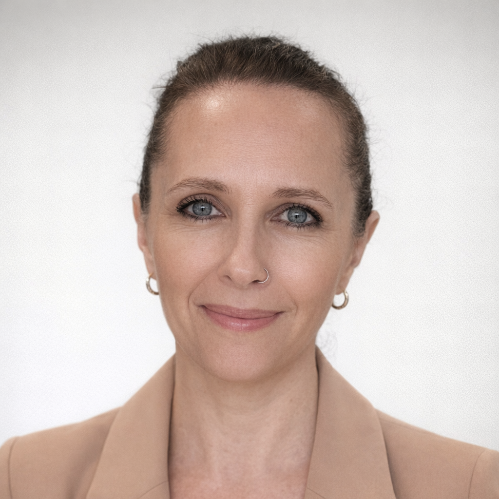 A woman with blue eyes, light makeup, and a slight smile, wearing gold hoop earrings and a tan blazer, with her hair pulled back, posing against a plain white background.
