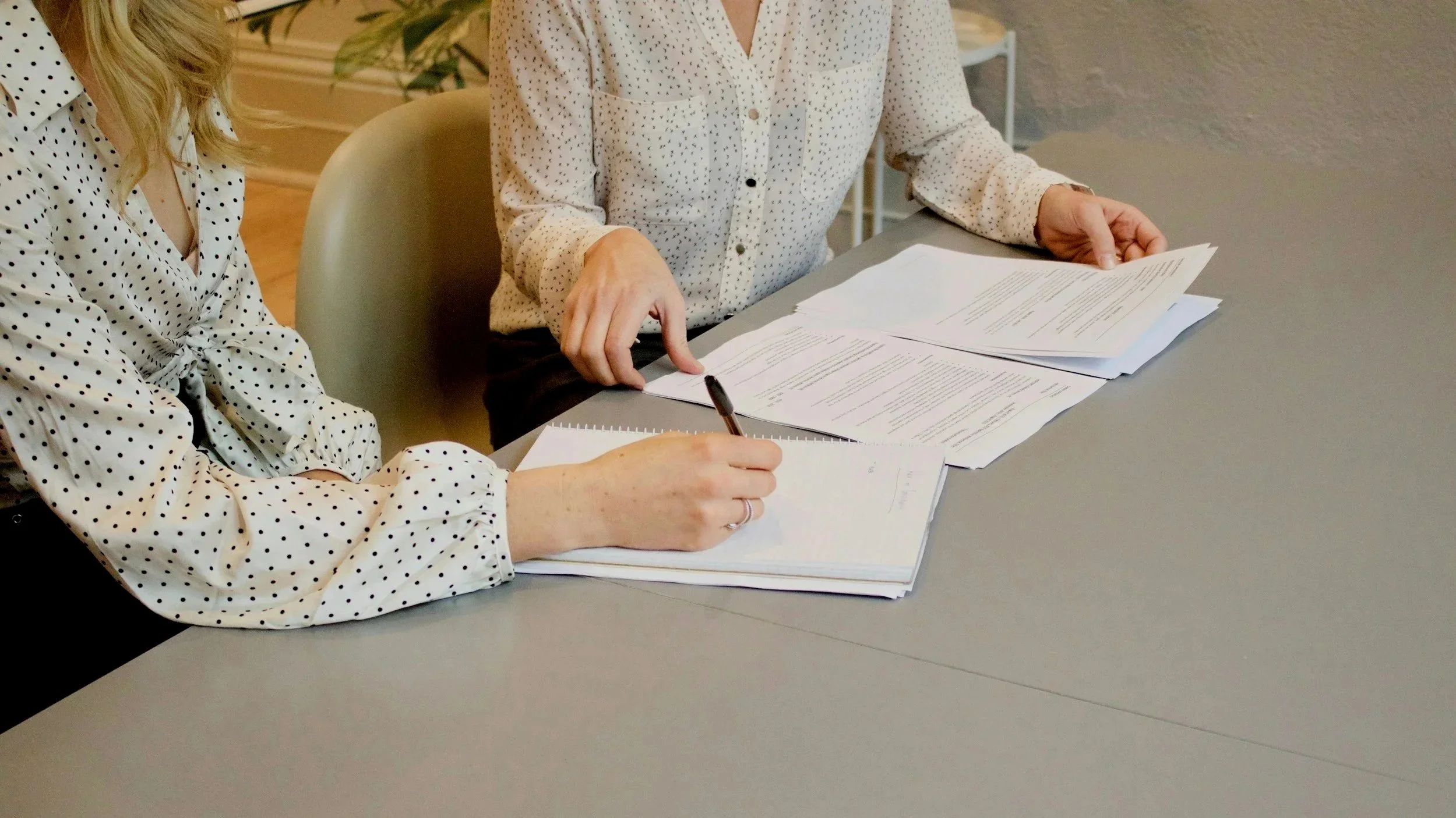 Two women wearing polka dot shirts sitting at a table with papers, documents, and a pen, engaged in a discussion about the documents.