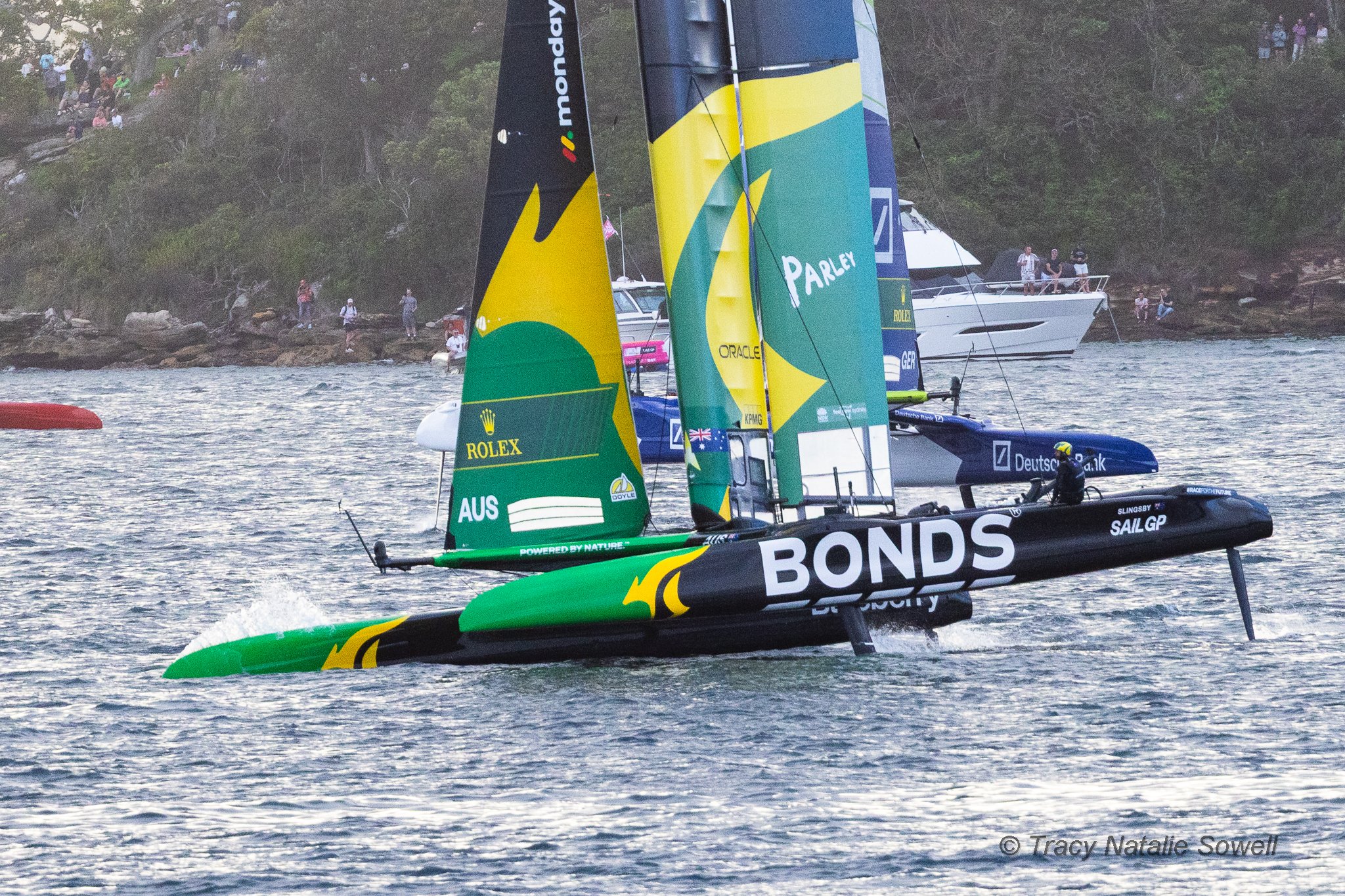 A scene of multiple racing sailboats on the water, with the sails displaying sponsors' logos like Bonds and Rolex. In the background, there are spectators on the shore, some standing and some sitting among trees and rocks.