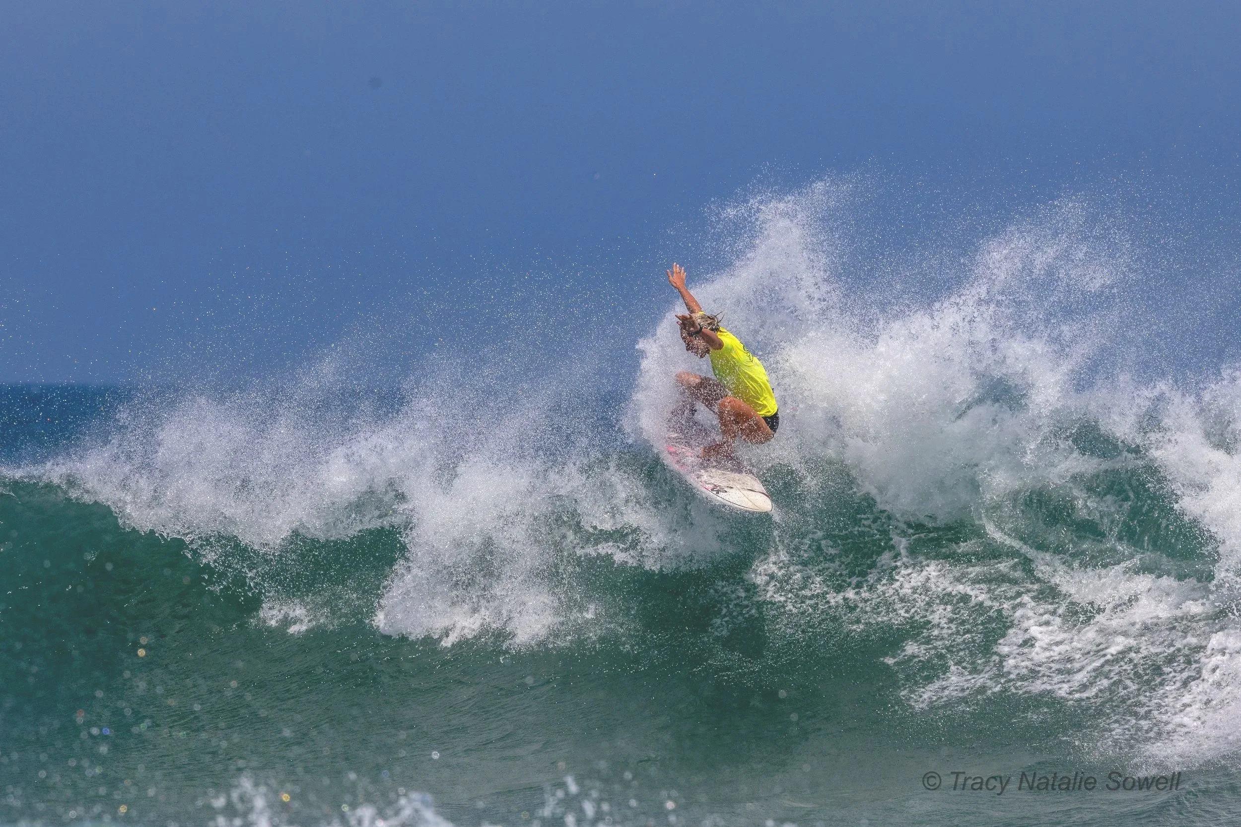 A person surfing on a wave in the ocean, wearing a yellow shirt and black shorts.