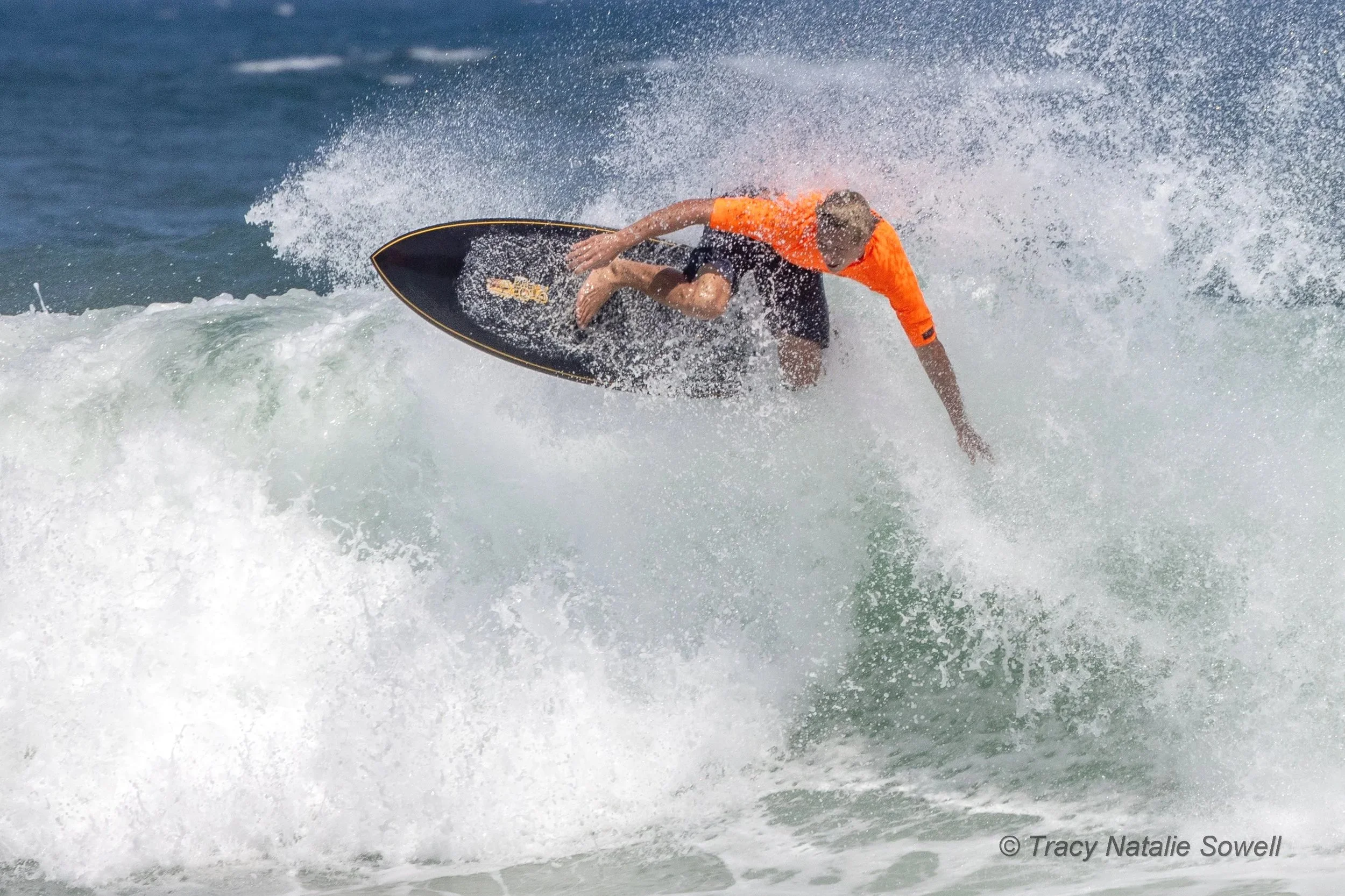 A person wearing an orange shirt and black shorts riding a surfboard on a wave, leaning into the wave with water spraying around.