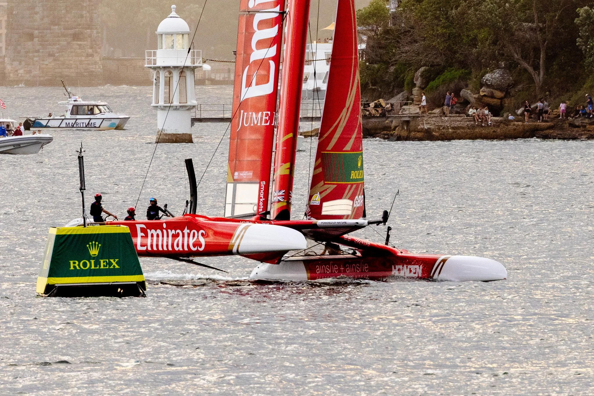 A racing sailboat with red sails featuring sponsorship logos from Emirates and Rolex, floating on a body of water with several crew members onboard, and a small pursuit boat nearby. In the background, there is a rocky shoreline with people sitting an