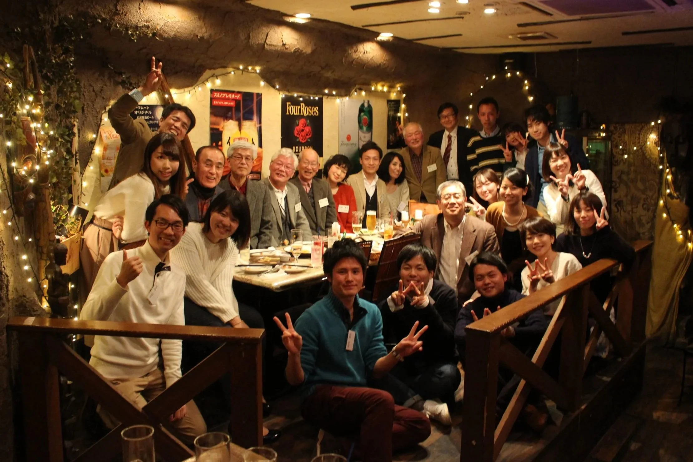 飲み会のグループ写真、多くの人が笑顔でポーズやピースサインをしている夜のレストランや居酒屋の室内。背景にライトとポスターが見える。