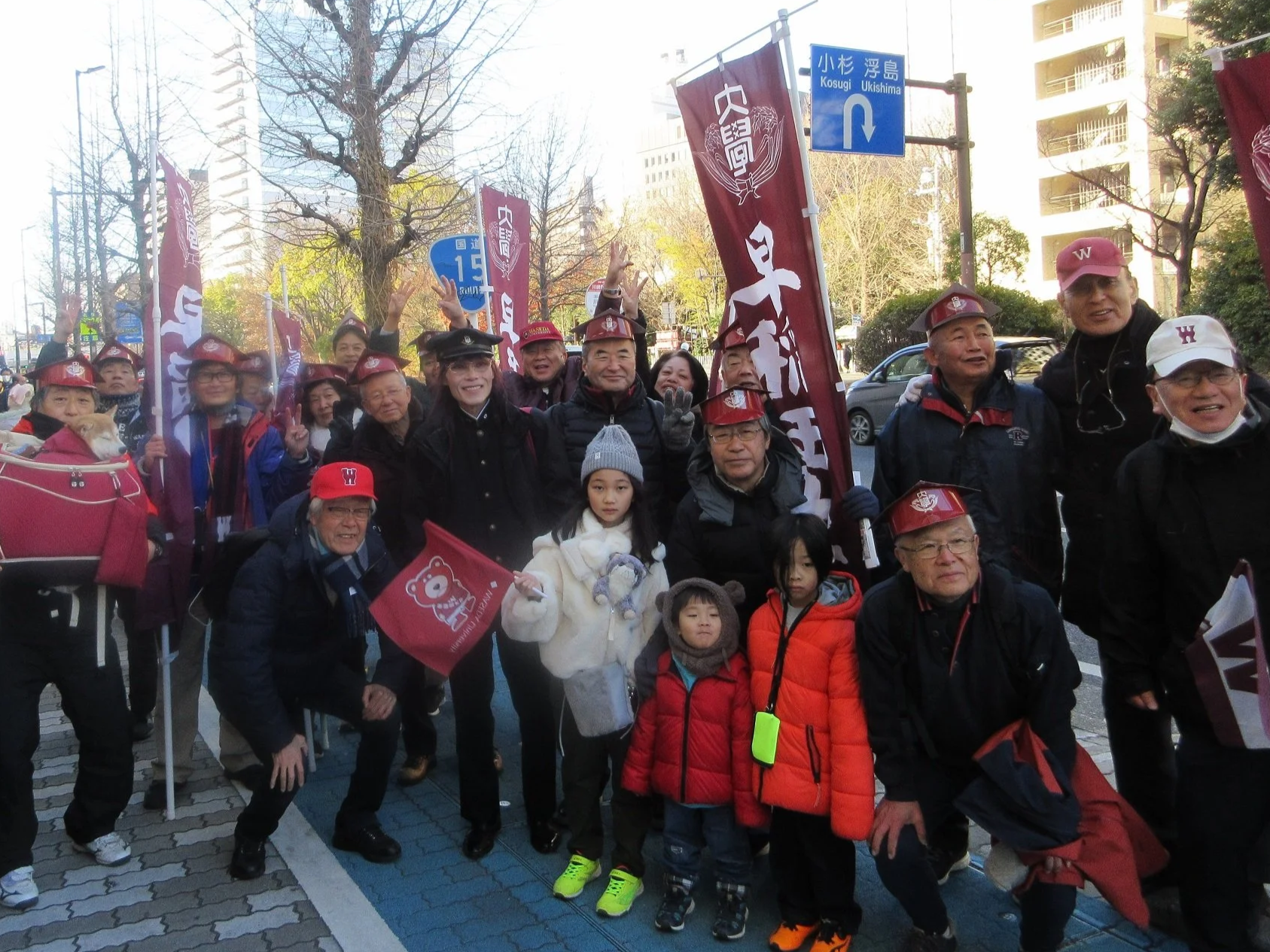 東京の街角で多くの人々が集まるイベントのグループ写真。みんな帽子やジャケットを着ており、いくつかの旗やバナーを持っている。背景には木やビルが見える。