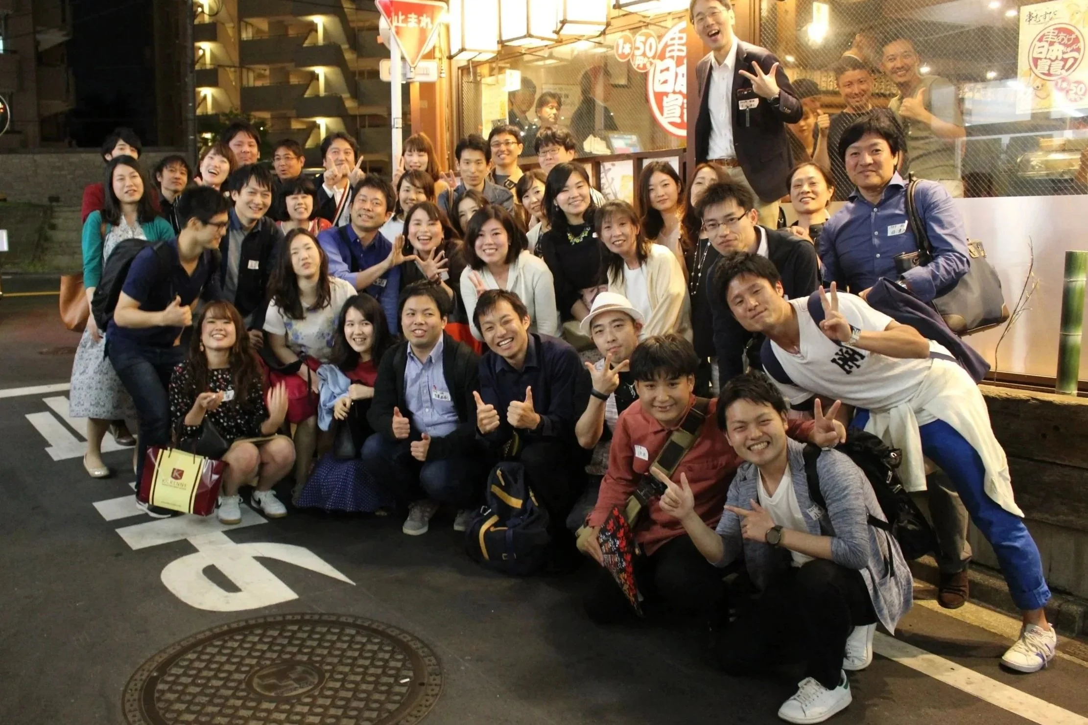 夜の街で撮影された、多数の笑顔のグループ写真。背景には商店や案内表示が見える。