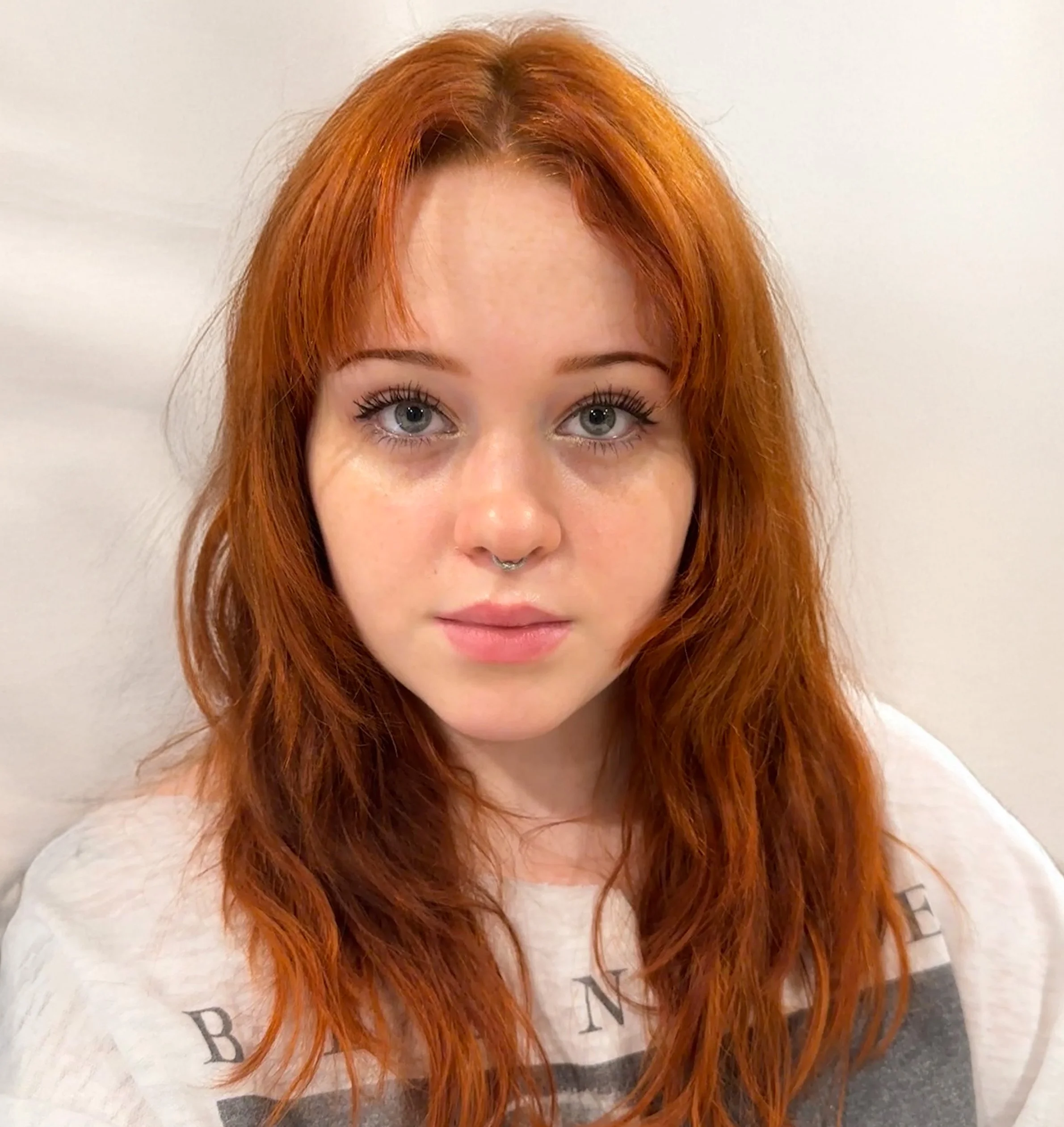 Close-up of a woman with bright red hair, blue eyes, and a septum piercing, wearing a white shirt.