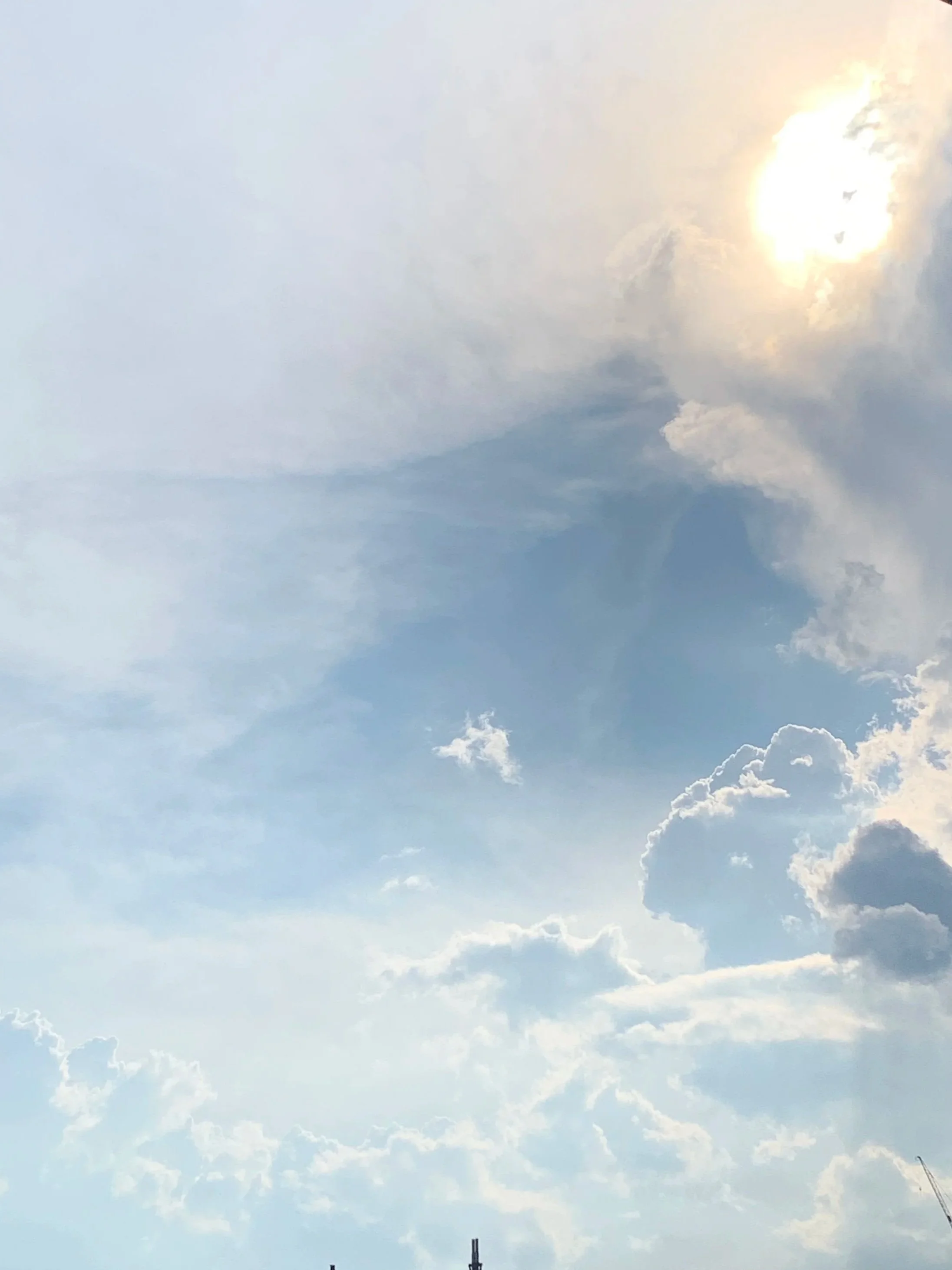 The sky with mostly light clouds and the sun partially obscured, shining brightly with a few darker clouds at the bottom.