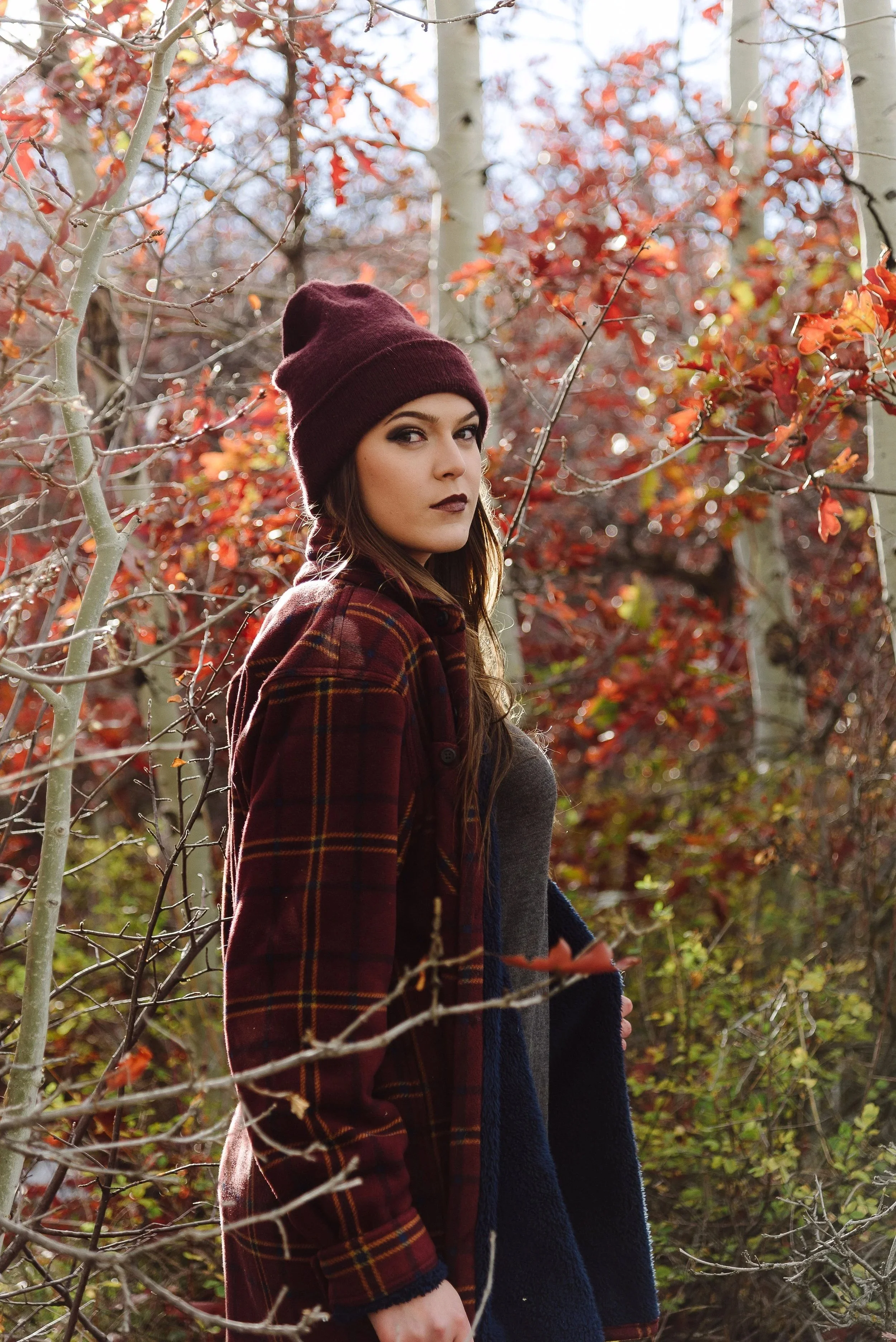 A woman with long dark hair wearing a dark red knit beanie and dark makeup stands outdoors among leafless trees with red and orange autumn leaves, looking at the camera.