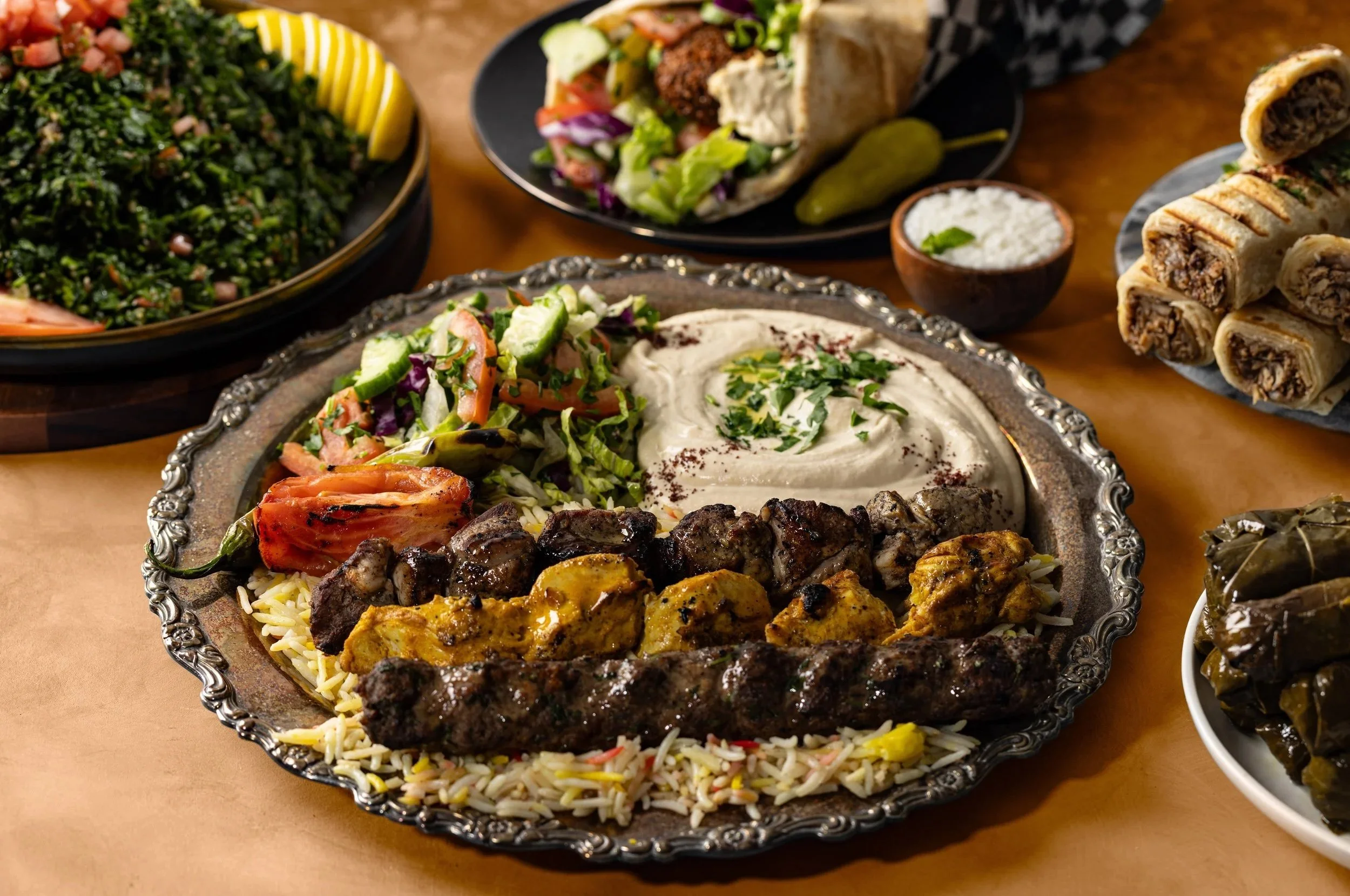 An assortment of Middle Eastern dishes including grilled meats, rice pilaf, salad, hummus, stuffed grape leaves, and flatbread on serving plates.