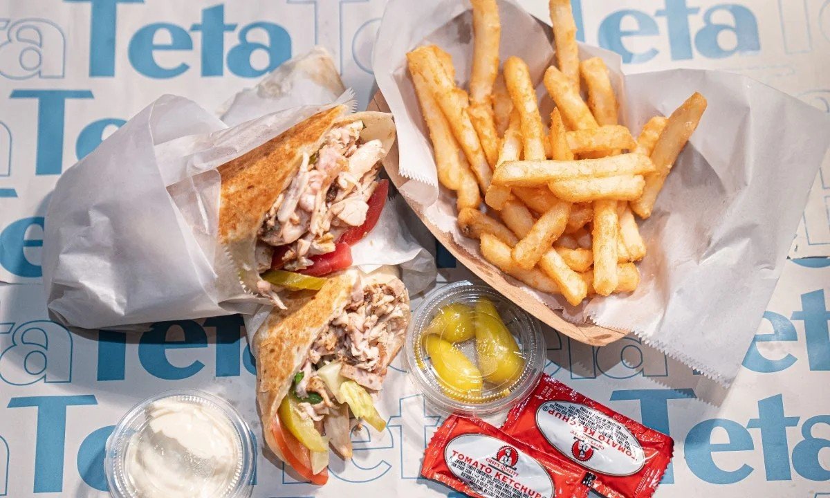 A meal with a chicken wrap, French fries, pickles, ketchup, and a small container of ranch dressing on a blue and white branded paper.
