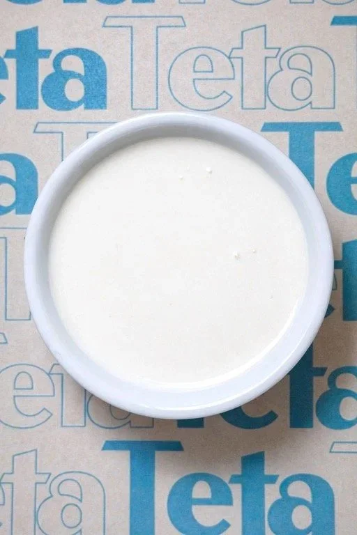 A small white bowl of sour cream on a surface with blue and white text that says "Teta" multiple times.