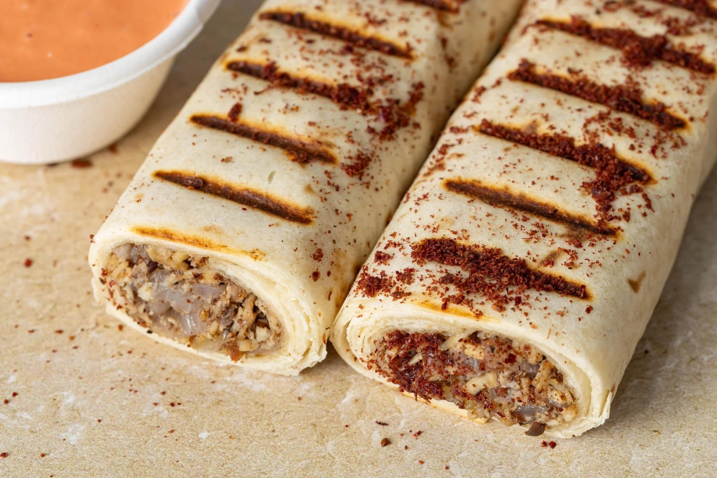 Close-up of two unwrapped ice cream burritos on a beige surface, with a cup of orange sauce or dip partially visible in the upper left corner.
